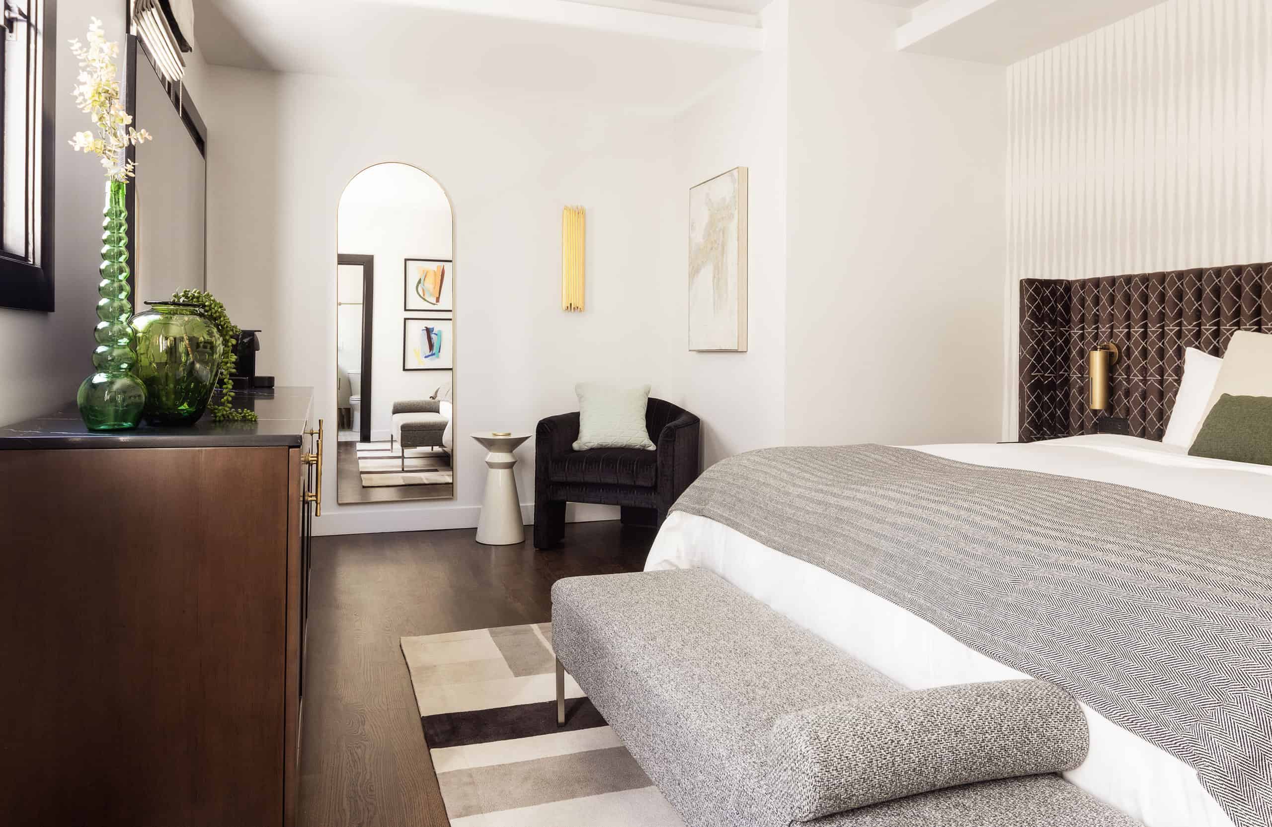 Minimalist bedroom with upholstered bed, neutral bedding, modern artwork, and arched mirror reflecting seating area.