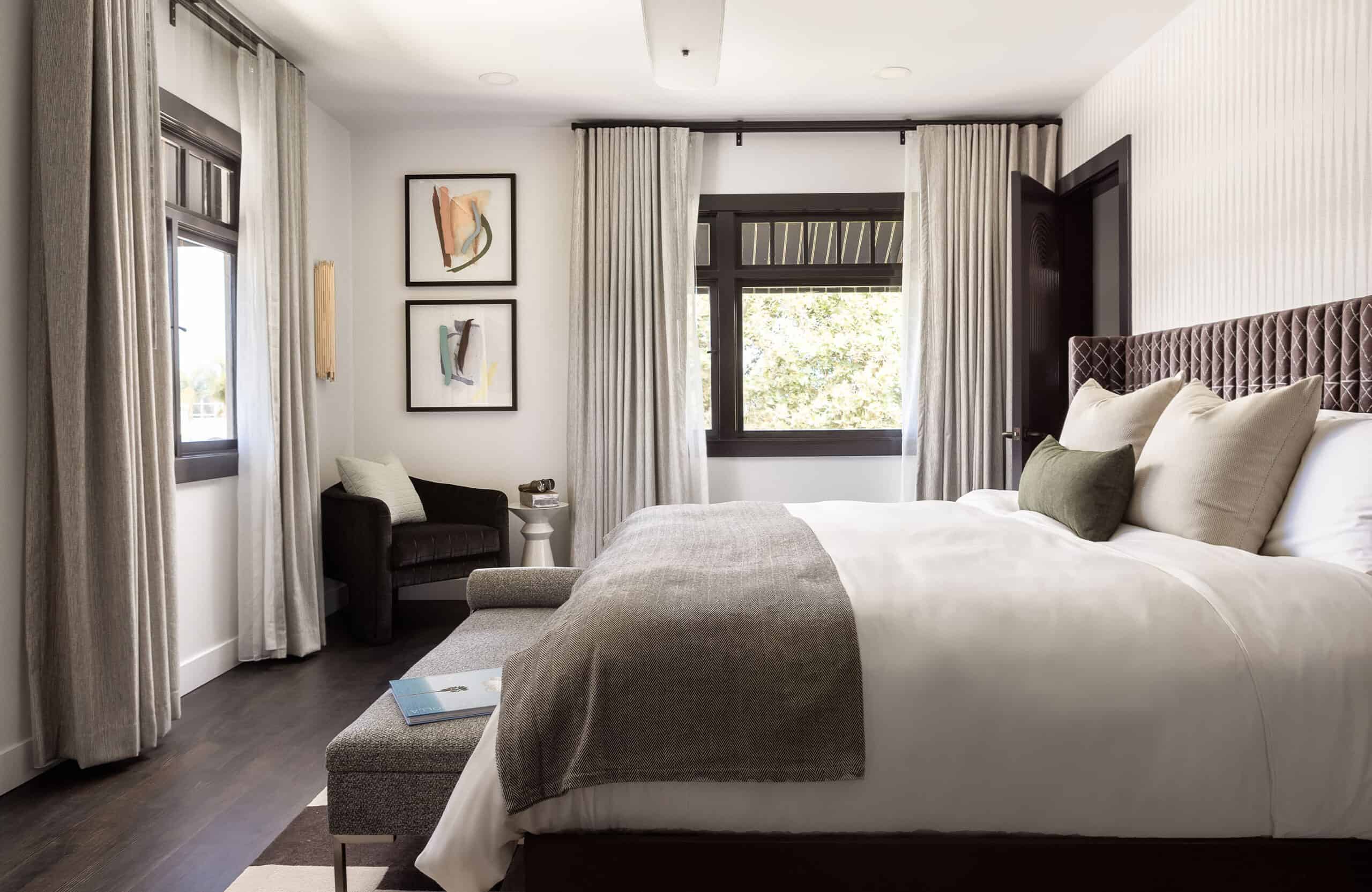 Bright hotel bedroom with a king bed, upholstered bench, soft gray textiles, and large windows.