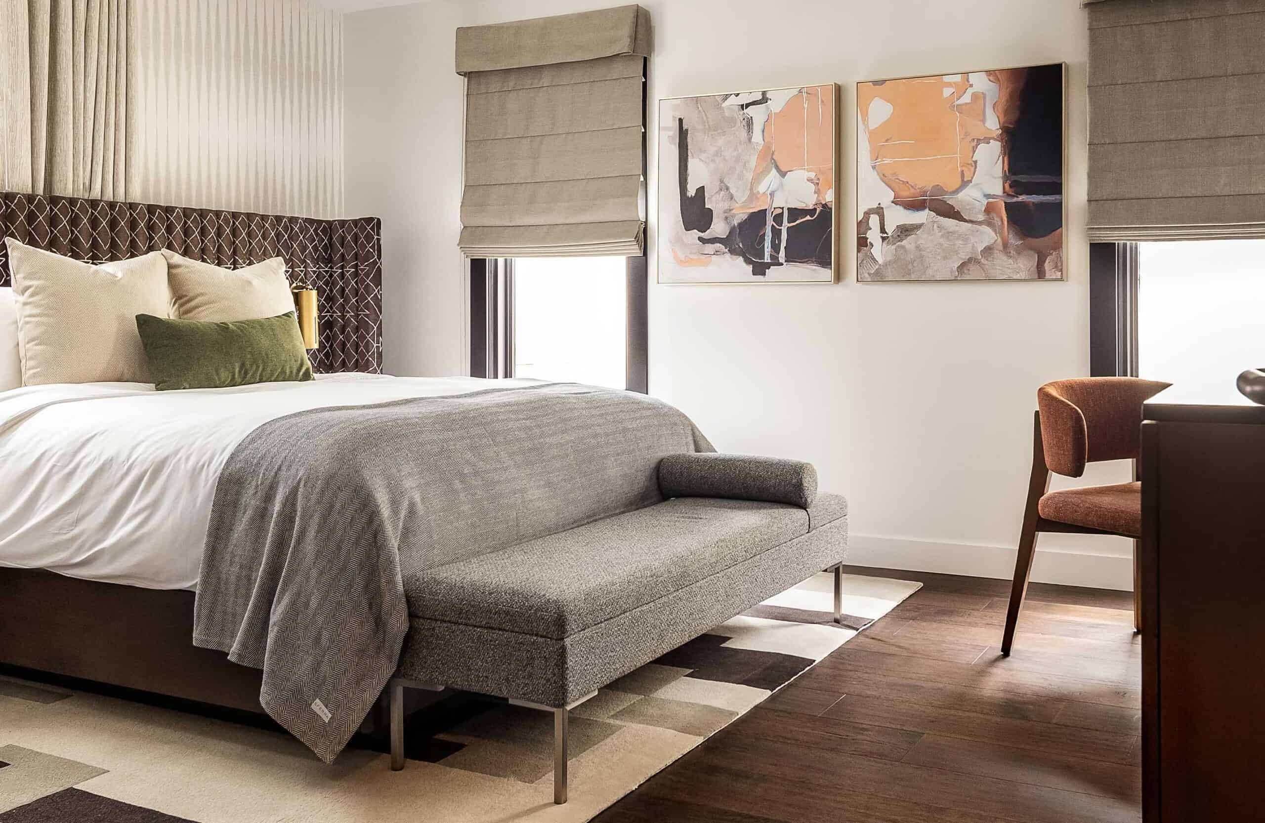 Sophisticated hotel bedroom with king bed, textured headboard, upholstered bench, abstract wall art, and warm wood flooring.