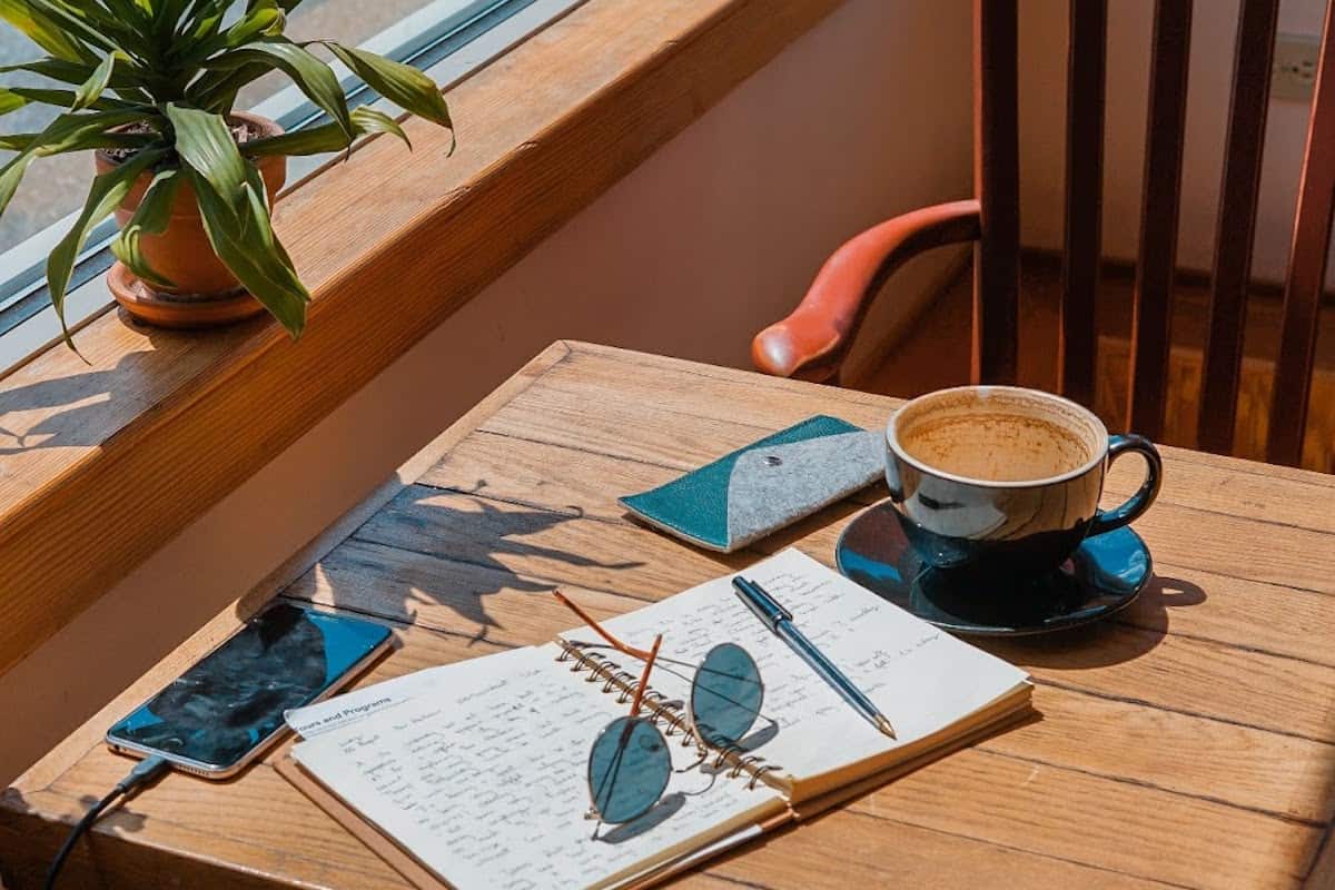 Sunlit café table with open notebook, sunglasses, smartphone, and a cup of coffee by a window