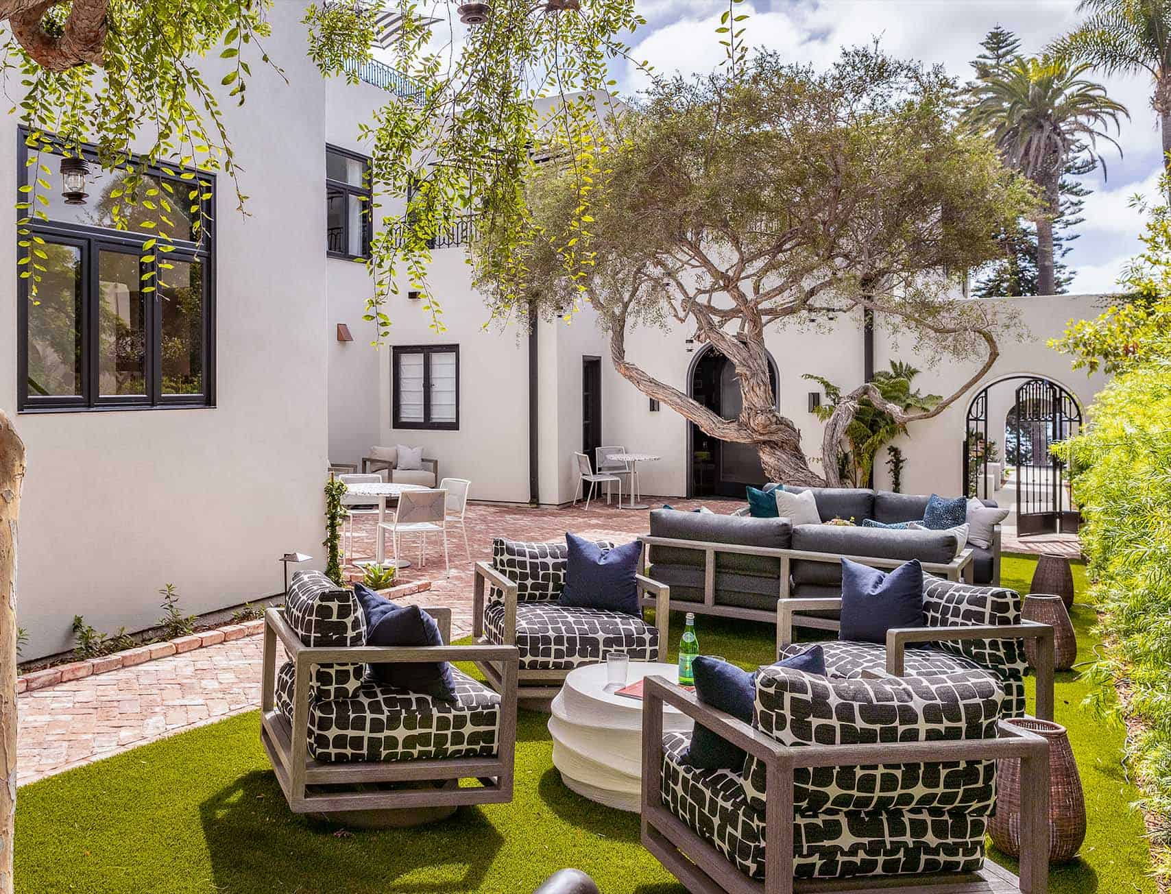 Outdoor courtyard seating area with patterned lounge furniture, umbrellas, and lush trees beside a white Mediterranean-style building