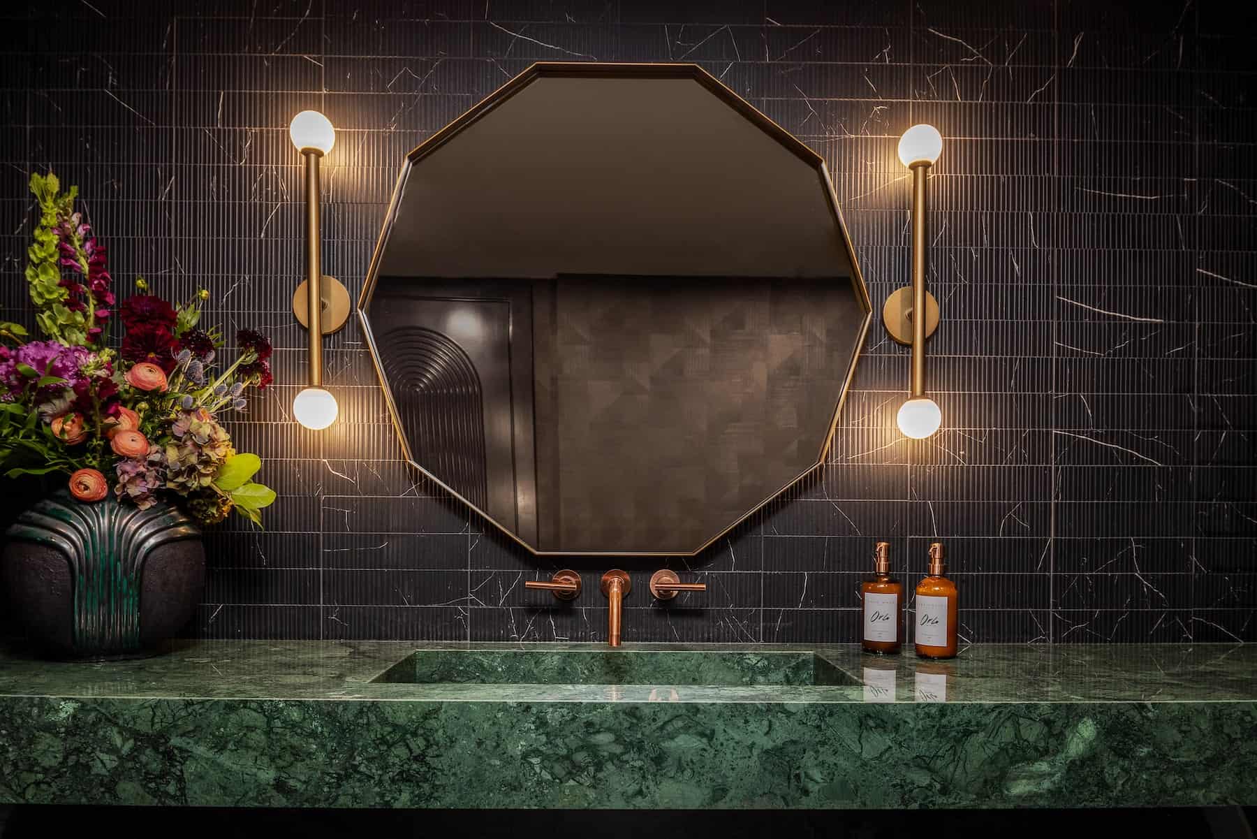Luxury bathroom vanity with green marble counter, brass fixtures, octagonal mirror, and modern wall lighting.