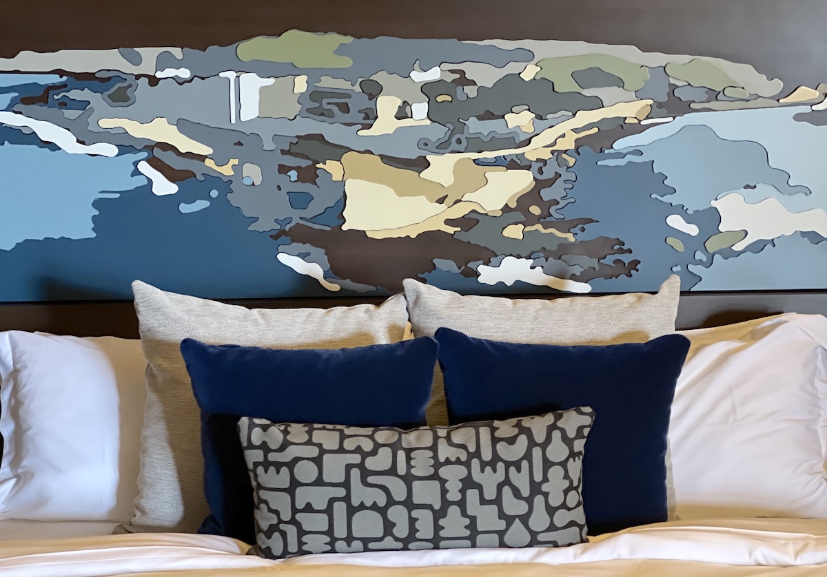 Styled hotel bed with layered white and navy pillows beneath an abstract coastal-inspired wall art headboard