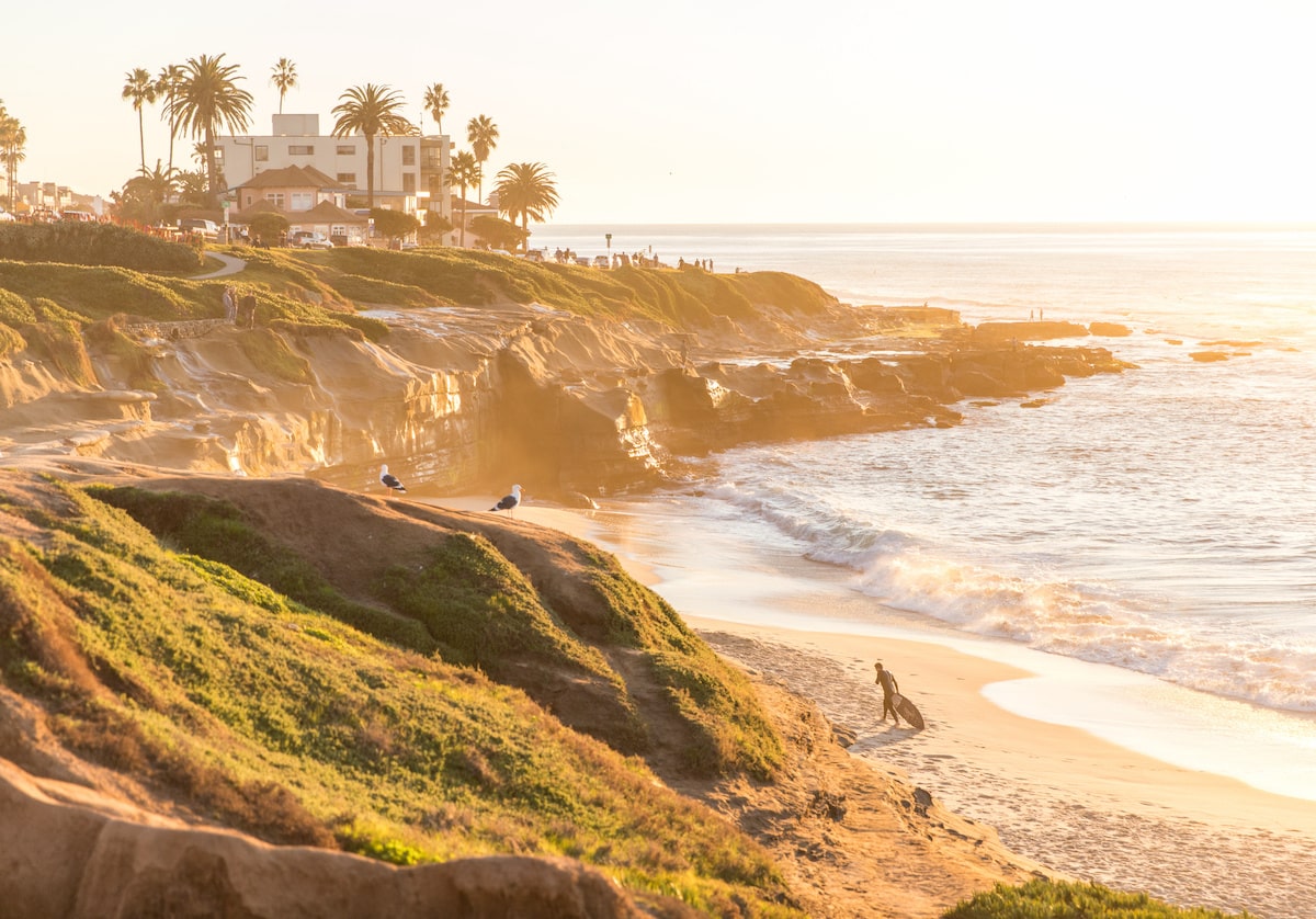 Golden sunset illuminating coastal cliffs and beach with palm trees, waves, and people walking along the shoreline.