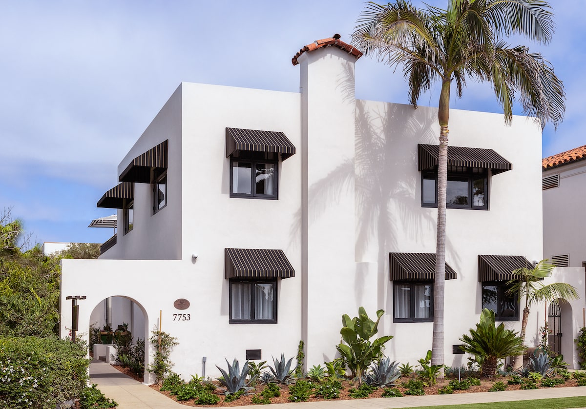 White Spanish-style two-story home with black awnings, palm tree landscaping, and arched entryway.