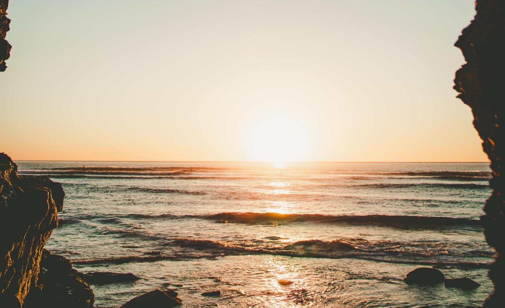 Golden sunset over the ocean viewed from a rocky coastal cove, with gentle waves reflecting warm light along the shoreline.