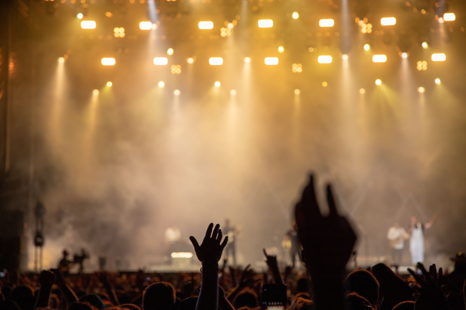 Audience raising hands at a live music concert with warm stage lighting and light haze.