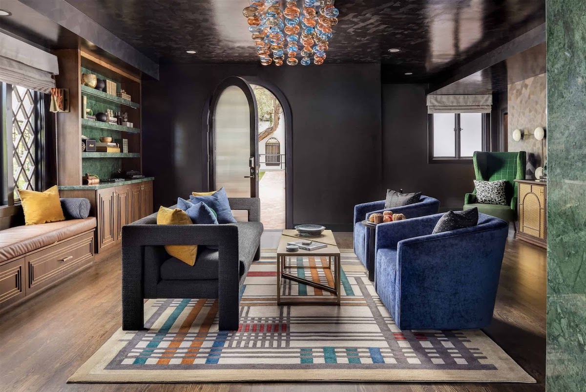 Moody boutique hotel lounge with modern seating, blue velvet armchairs, built-in shelving, patterned area rug, and an arched doorway leading to an outdoor courtyard.
