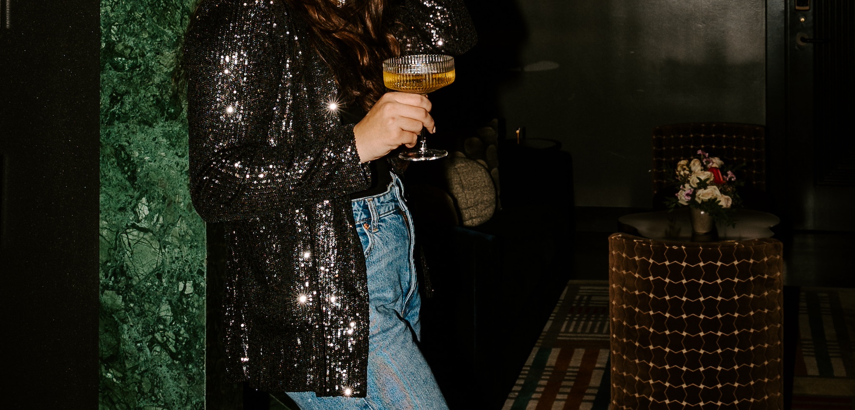 Person wearing a sequined jacket and jeans holding a cocktail in a dimly lit, stylish bar or lounge setting