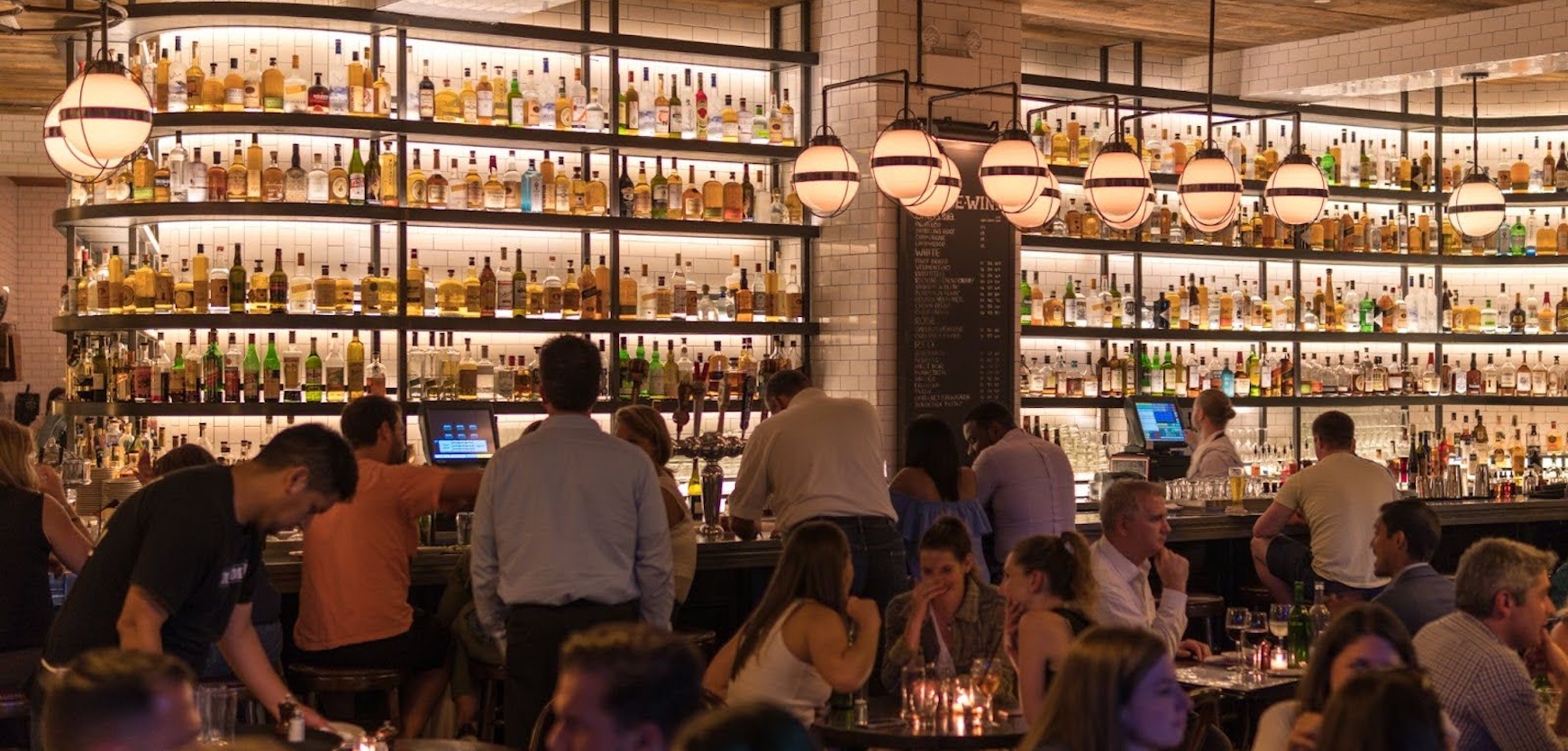 Lively upscale bar interior with guests socializing beneath shelves of illuminated liquor bottles and modern pendant lighting