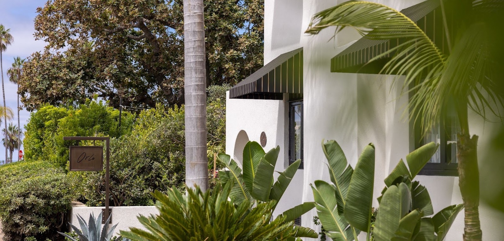 White stucco building with black awnings surrounded by palm trees and lush tropical landscaping in a coastal setting