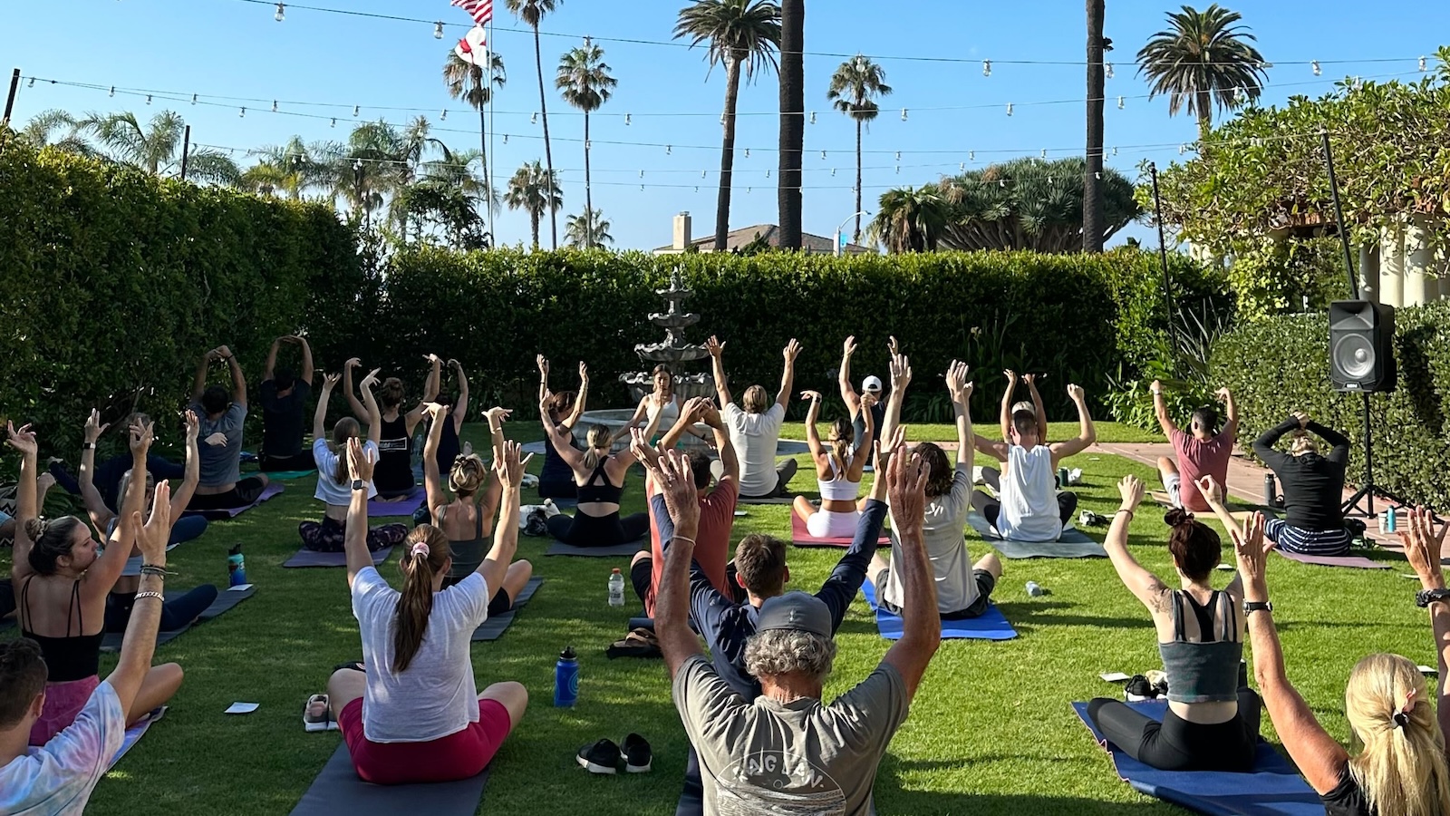 Outdoor group yoga class with participants seated on mats, raising their arms in a sunny garden surrounded by palm trees