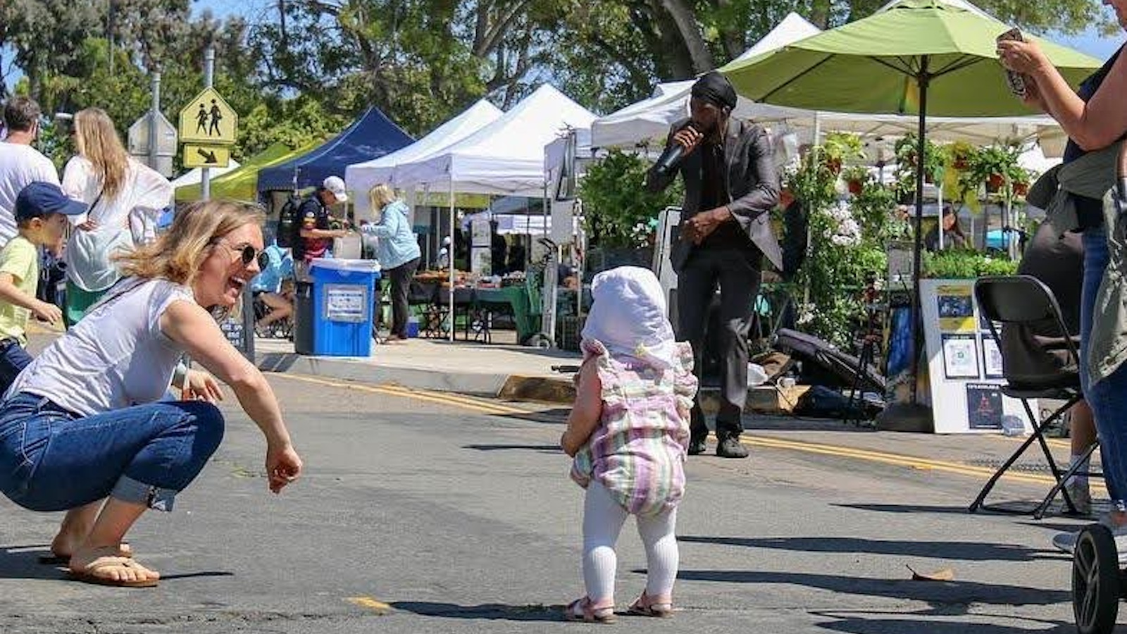 Woman crouching and smiling at a toddler walking at a lively outdoor community market with vendor tents and music in the background