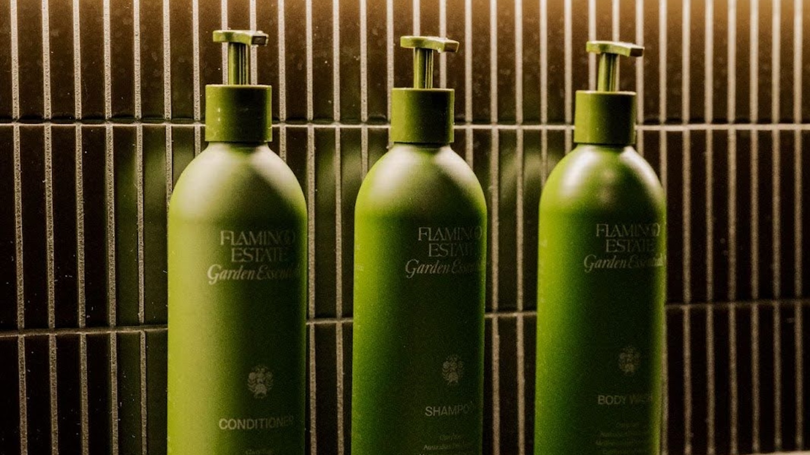 Green glass shampoo and conditioner bottles displayed against a dark tiled background in a luxury spa or hotel bathroom setting
