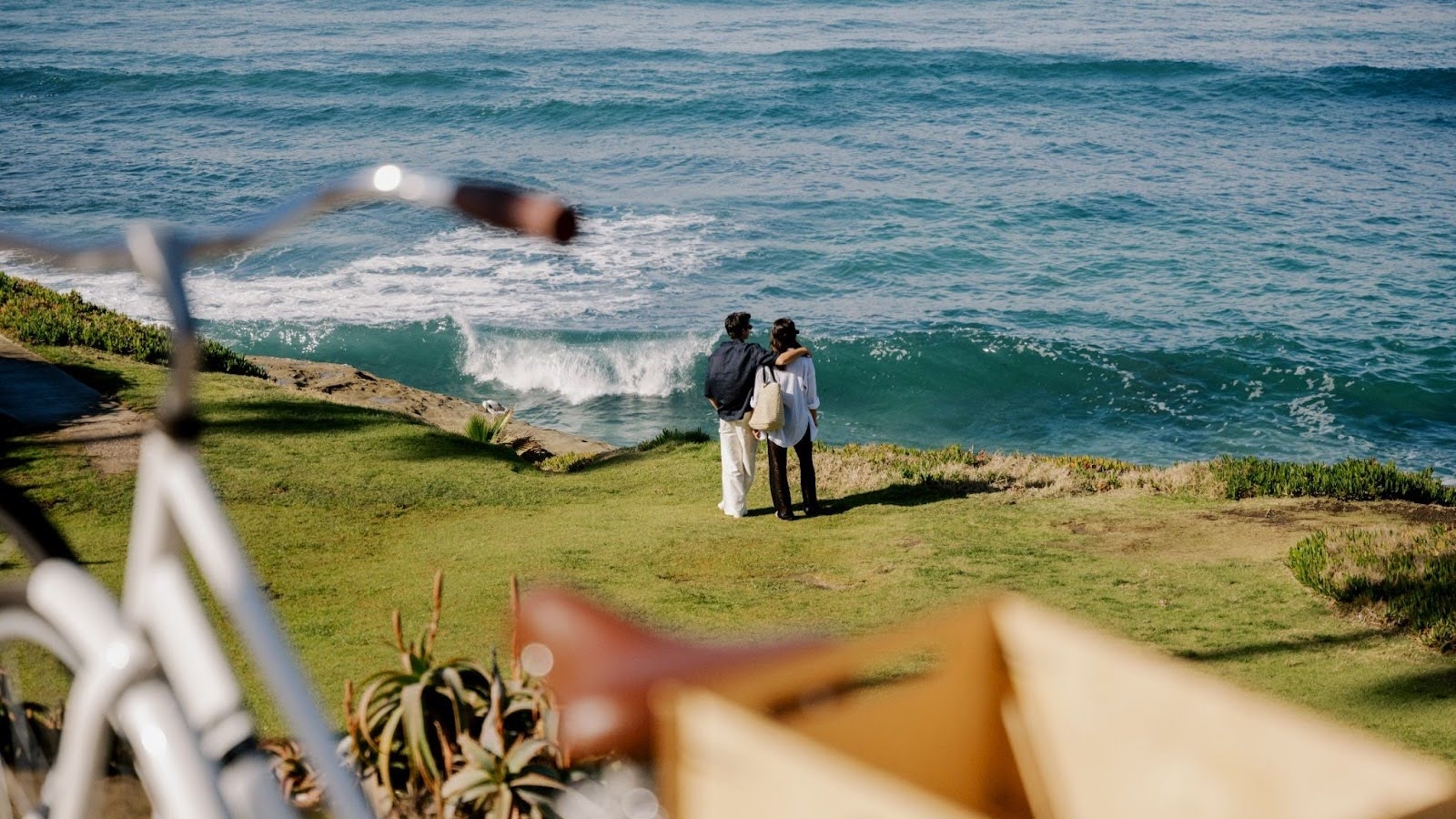 Couple standing on a grassy coastal bluff overlooking ocean waves, with bicycles in the foreground on a scenic seaside path