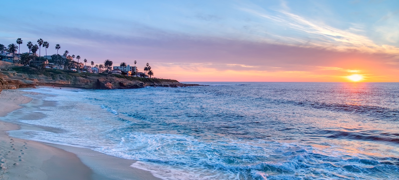 Sunset over a sandy beach with gentle waves, coastal cliffs, and palm trees along the shoreline