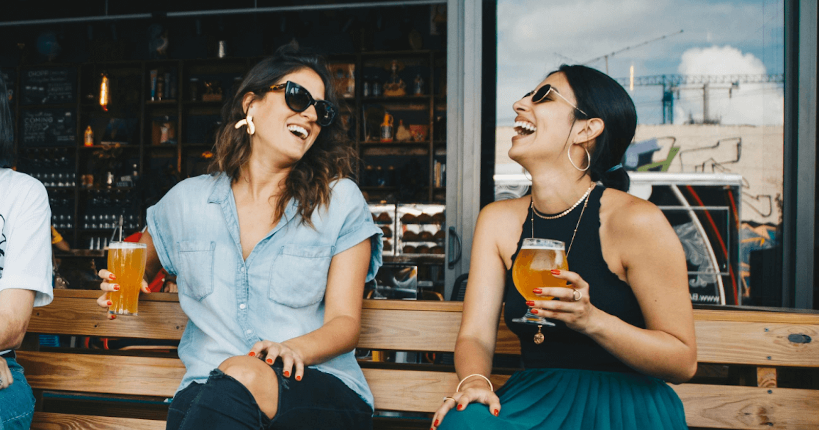 Two women laughing and enjoying craft beers while seated at an outdoor bar on a sunny day