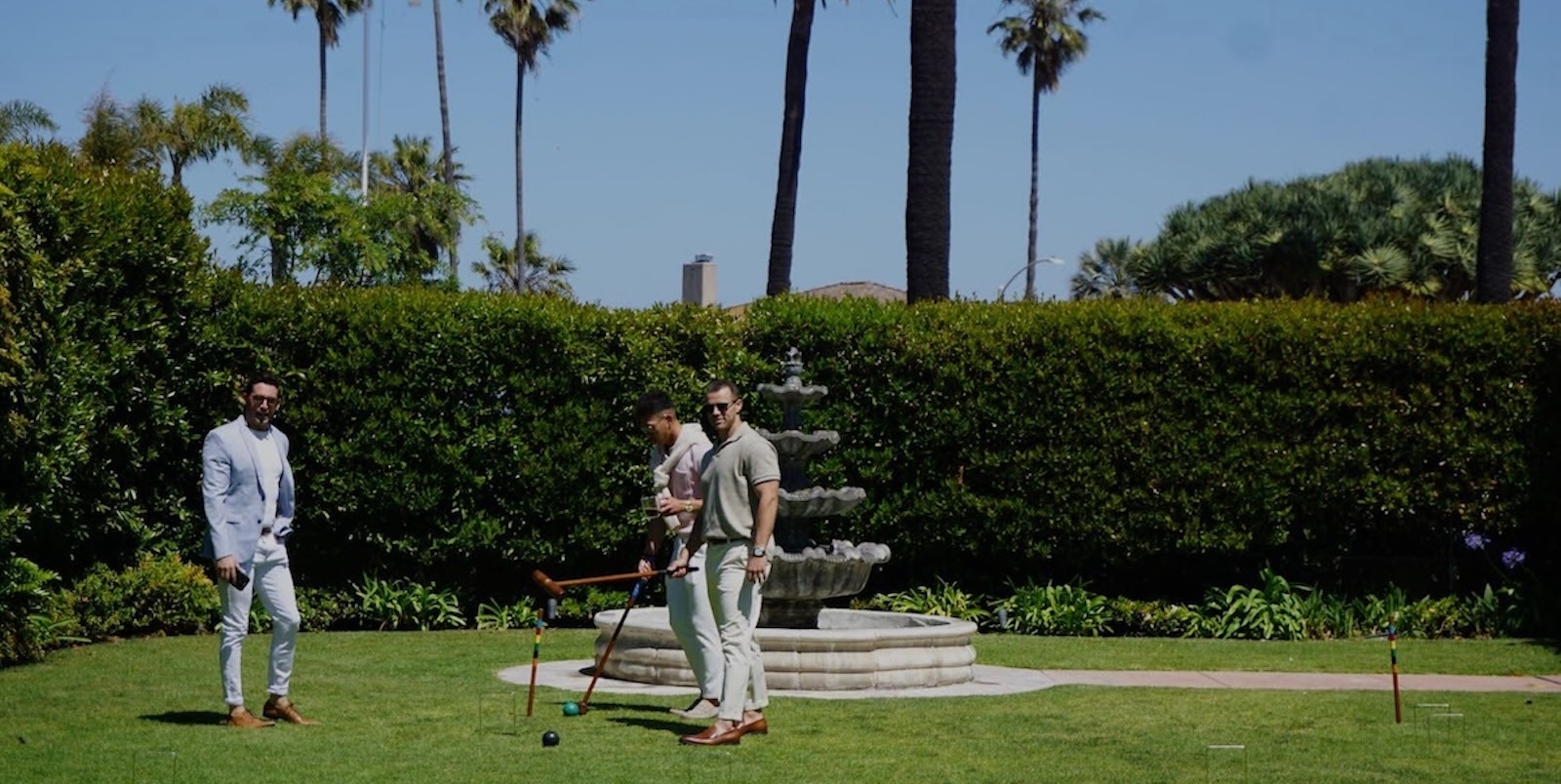 Three men playing croquet on a manicured lawn beside a stone fountain, dressed in light-colored casual outfits, with palm trees and hedges in a sunny garden setting.