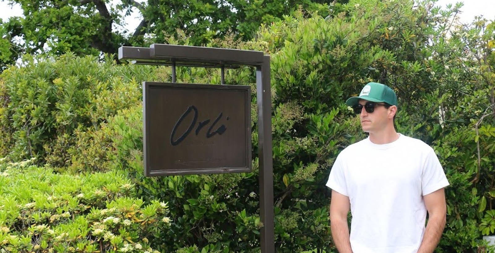 Man wearing a white t-shirt and green cap standing beside an “Orla” sign surrounded by lush greenery outdoors
