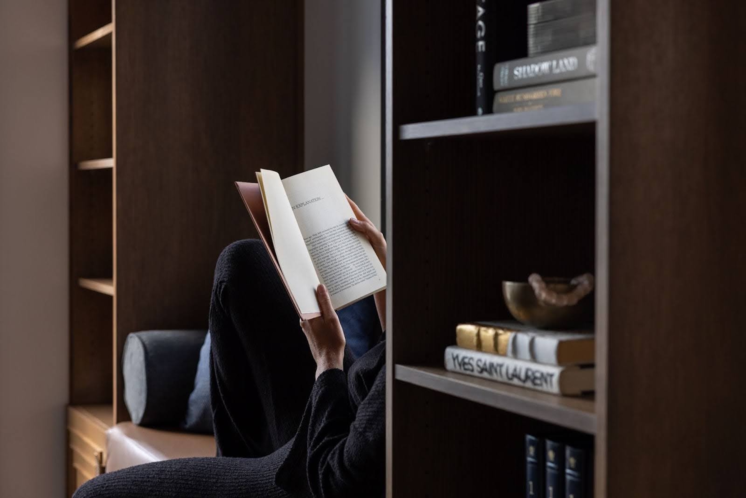 Person seated in a built-in reading nook holding an open book, surrounded by dark wood shelves and neatly arranged books in a quiet indoor space.