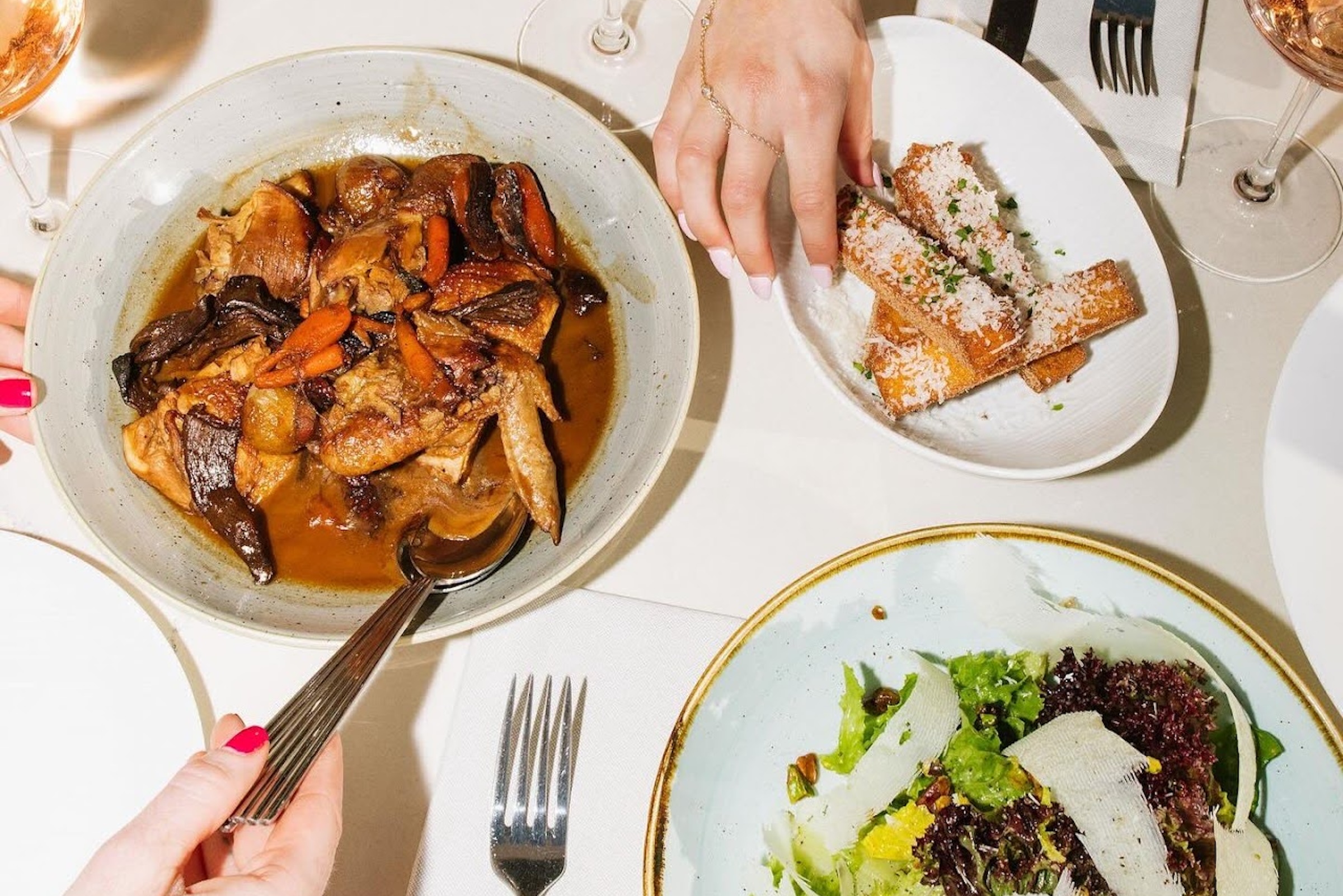 Overhead view of a shared restaurant table featuring braised chicken with mushrooms and carrots, crispy bread topped with cheese, and a fresh green salad, capturing an elevated communal dining experience.