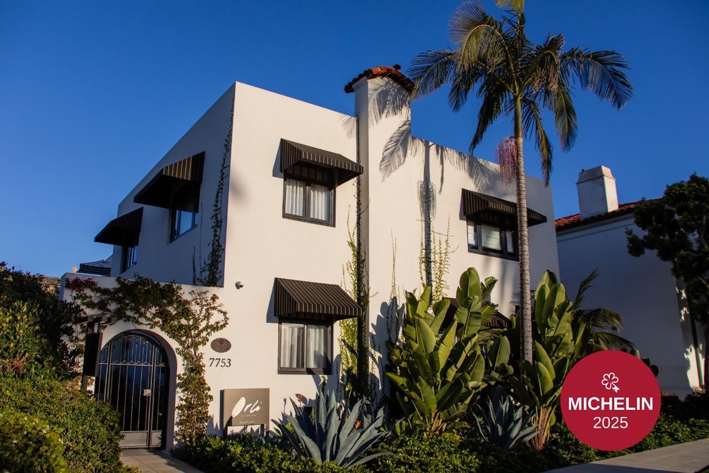 Exterior of a Michelin 2025–recognized boutique hotel with white stucco architecture, palm trees, and lush landscaping in a coastal California neighborhood.