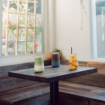 Three assorted drinks on a wooden table inside a bright café with window light and hanging plants