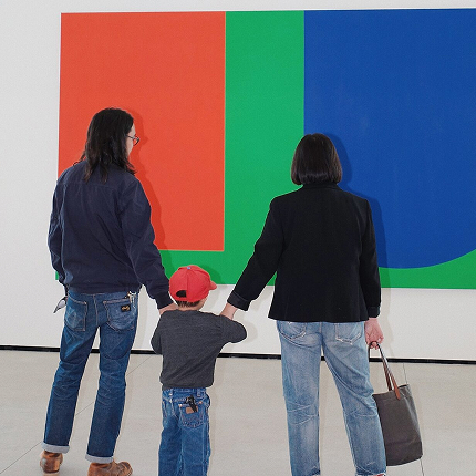 Two adults and a child holding hands while viewing a large abstract artwork with bold red, green, and blue shapes in a gallery