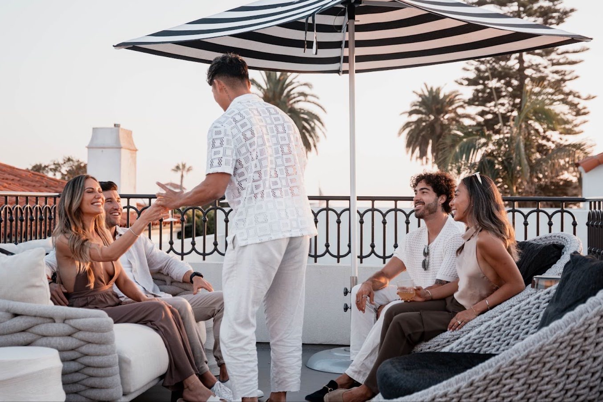 Friends gathered on a rooftop lounge at sunset, enjoying drinks and conversation under a striped patio umbrella.
