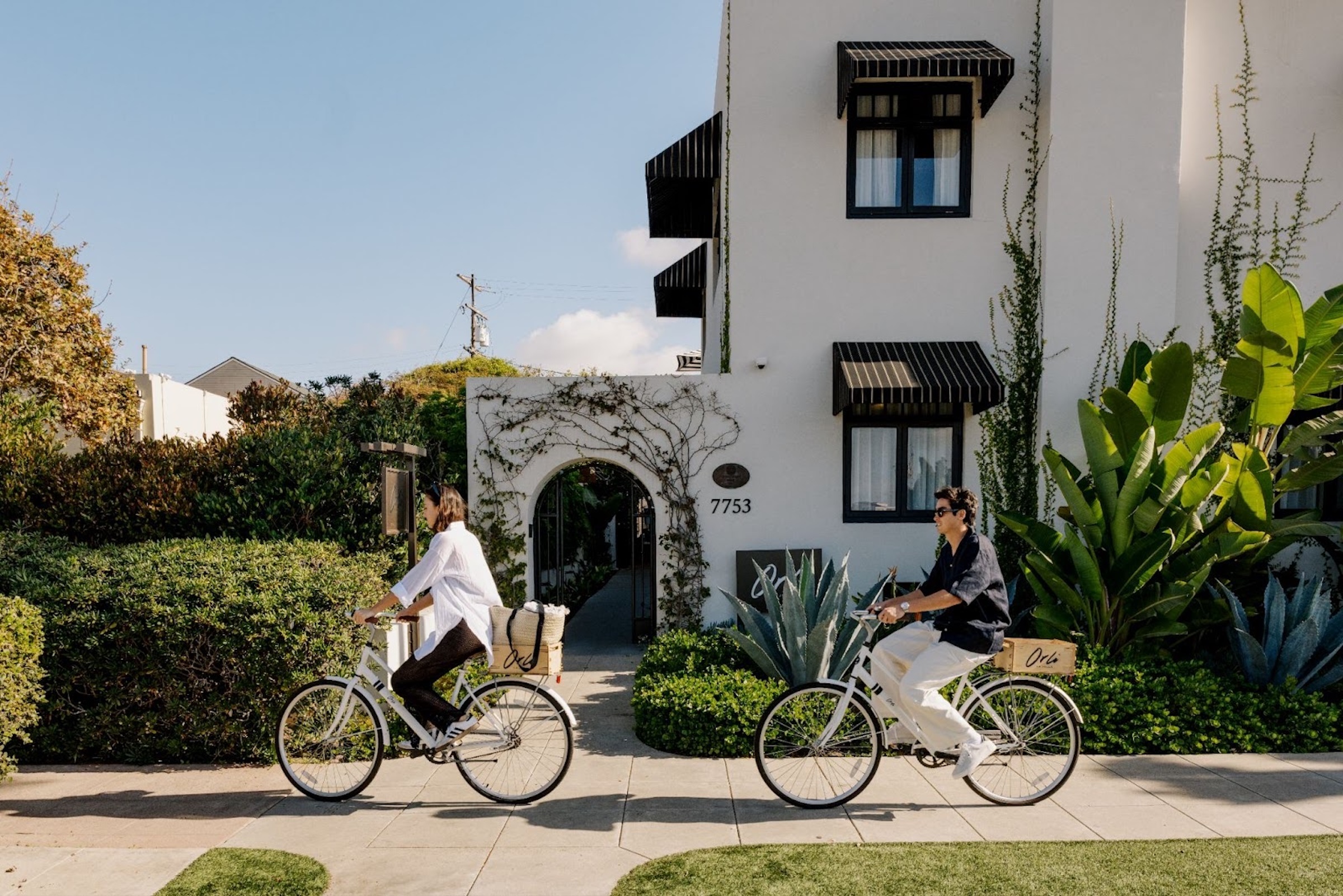 Two people riding bicycles past a white, ivy-covered building entrance on a sunny residential street.