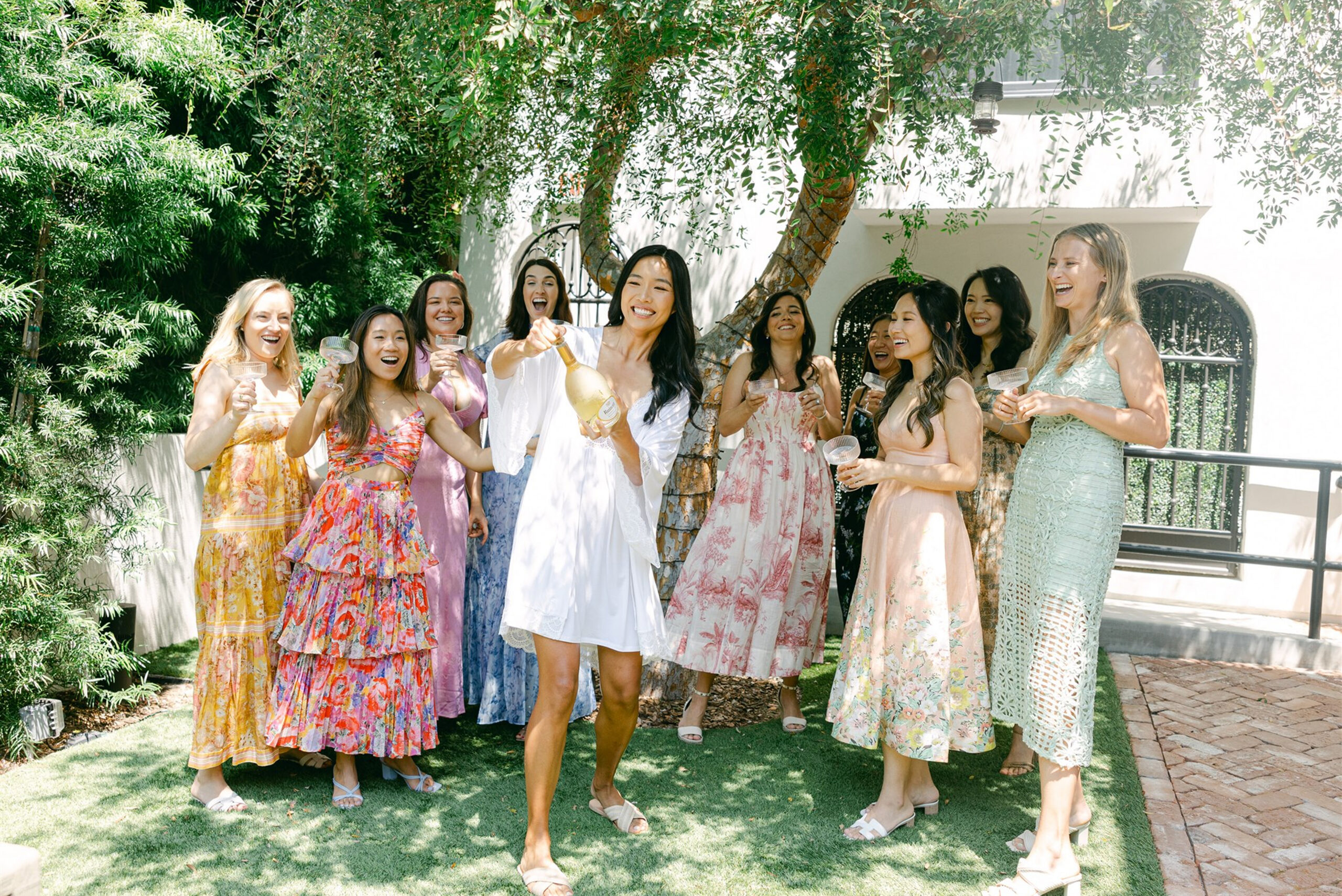 Bride popping a champagne bottle while surrounded by bridesmaids holding glasses and cheering in a sunlit garden courtyard.
