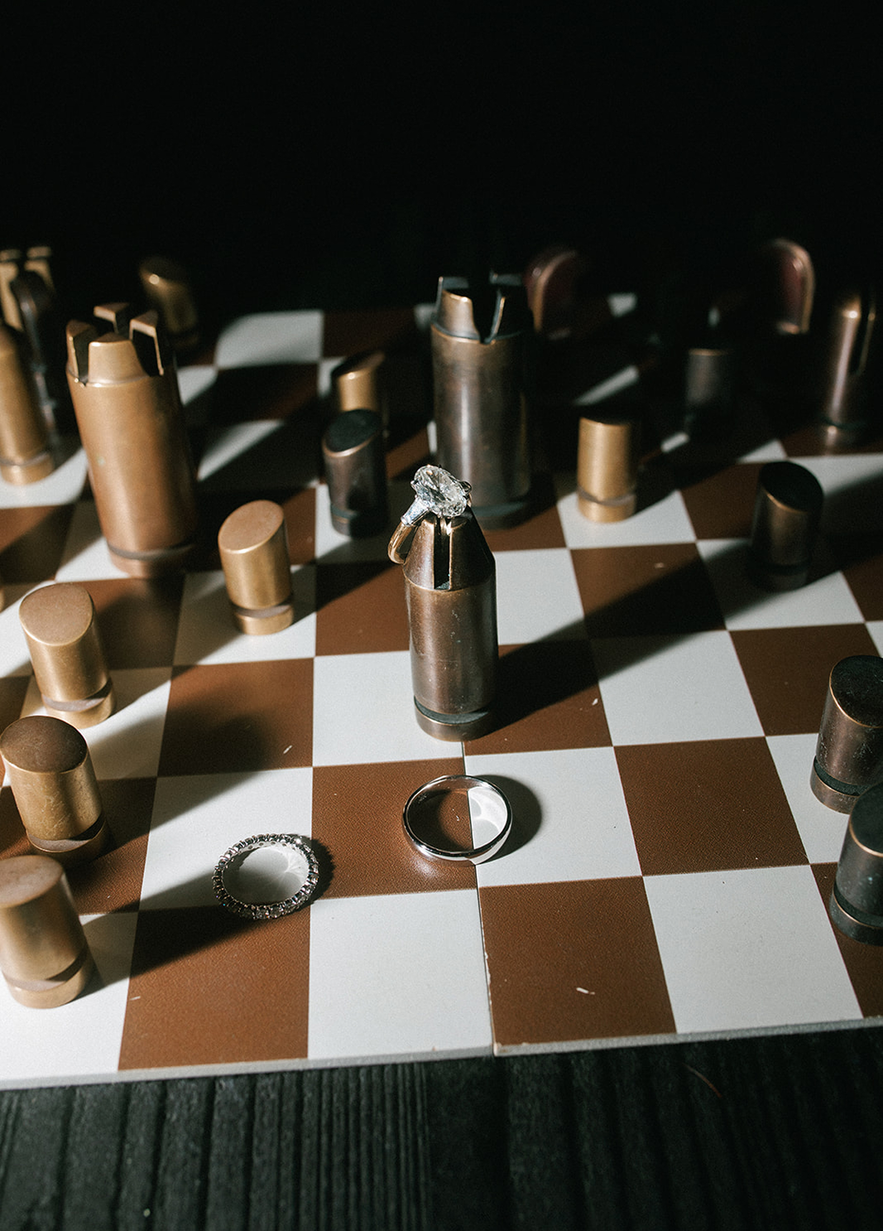 Wedding rings arranged on a chessboard with metallic chess pieces, featuring an engagement ring standing upright among the pieces.