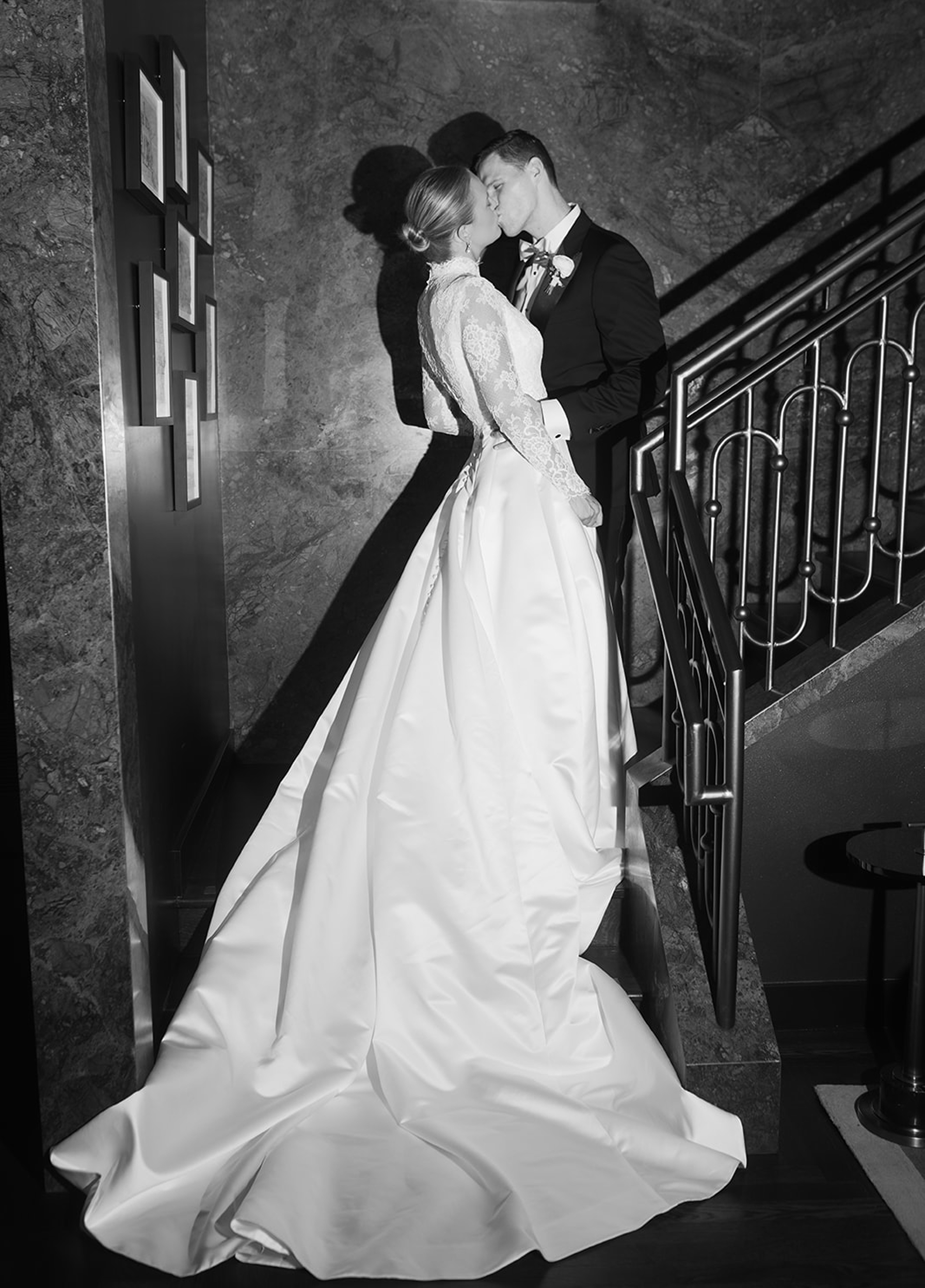 Black-and-white portrait of a bride and groom sharing a kiss on a staircase, with the bride’s long gown flowing behind her.