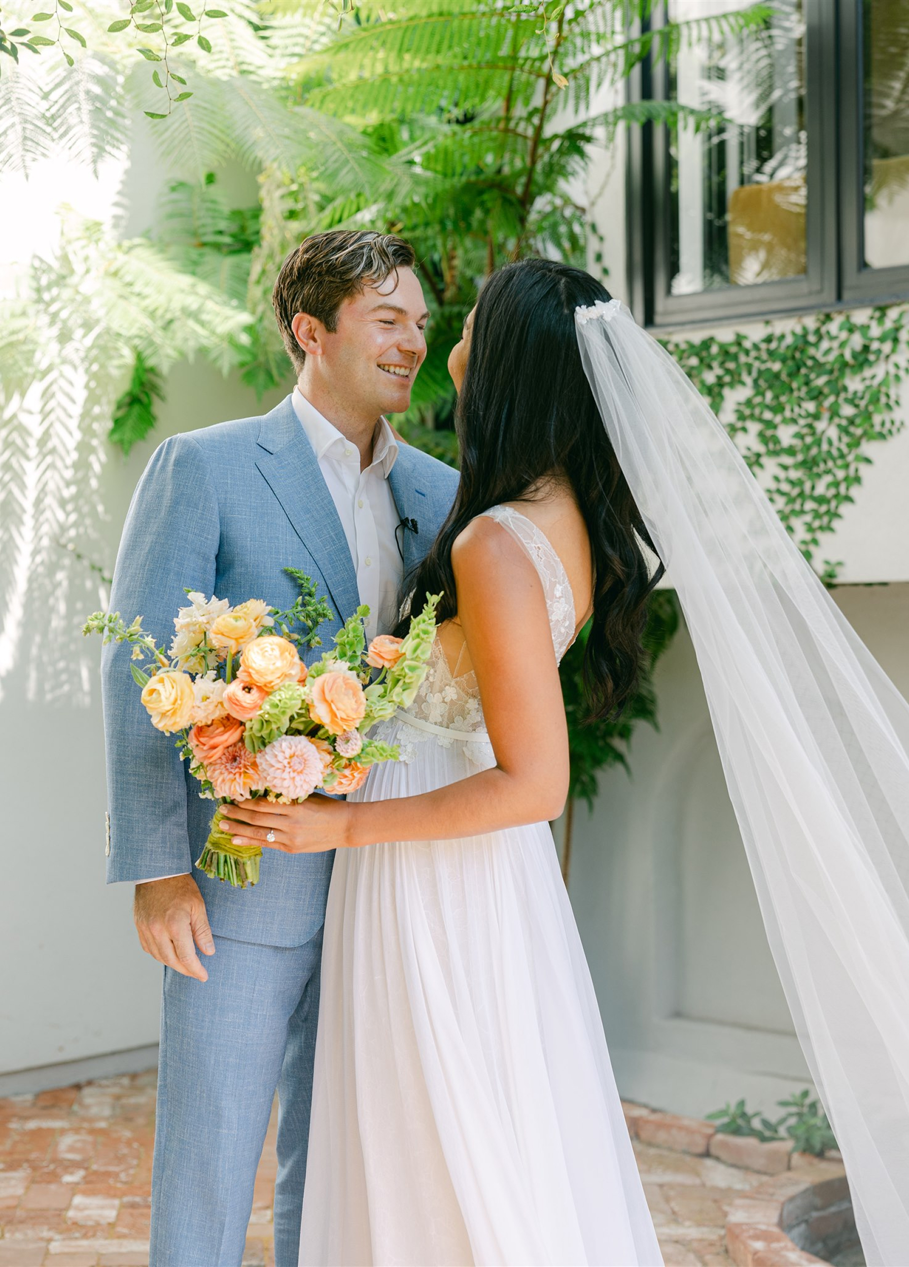 Bride holding a bouquet and smiling at the groom in a garden courtyard, with greenery behind them and the bride’s veil flowing over her shoulder.
