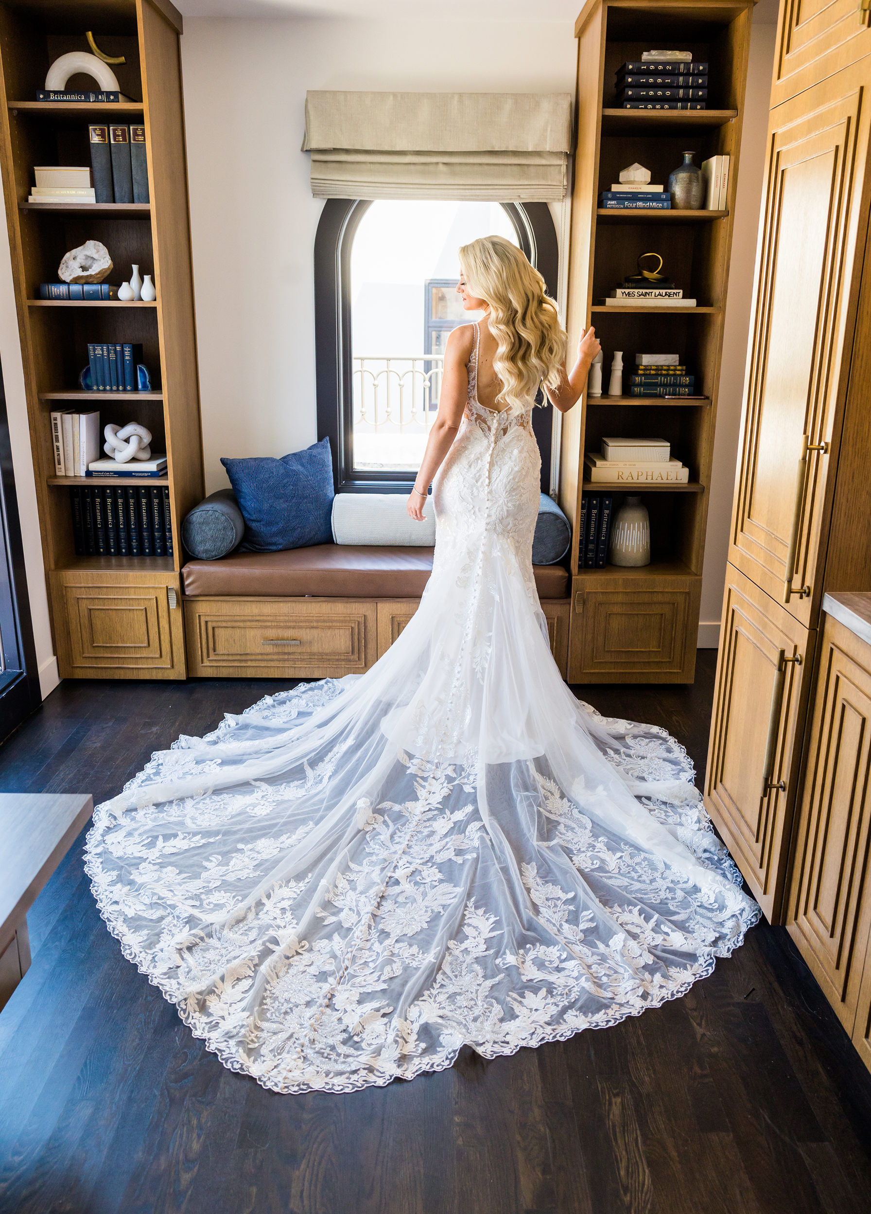 Bride standing indoors in a lace wedding gown with a long, detailed train flowing behind her, framed by built-in bookshelves and a window seat.