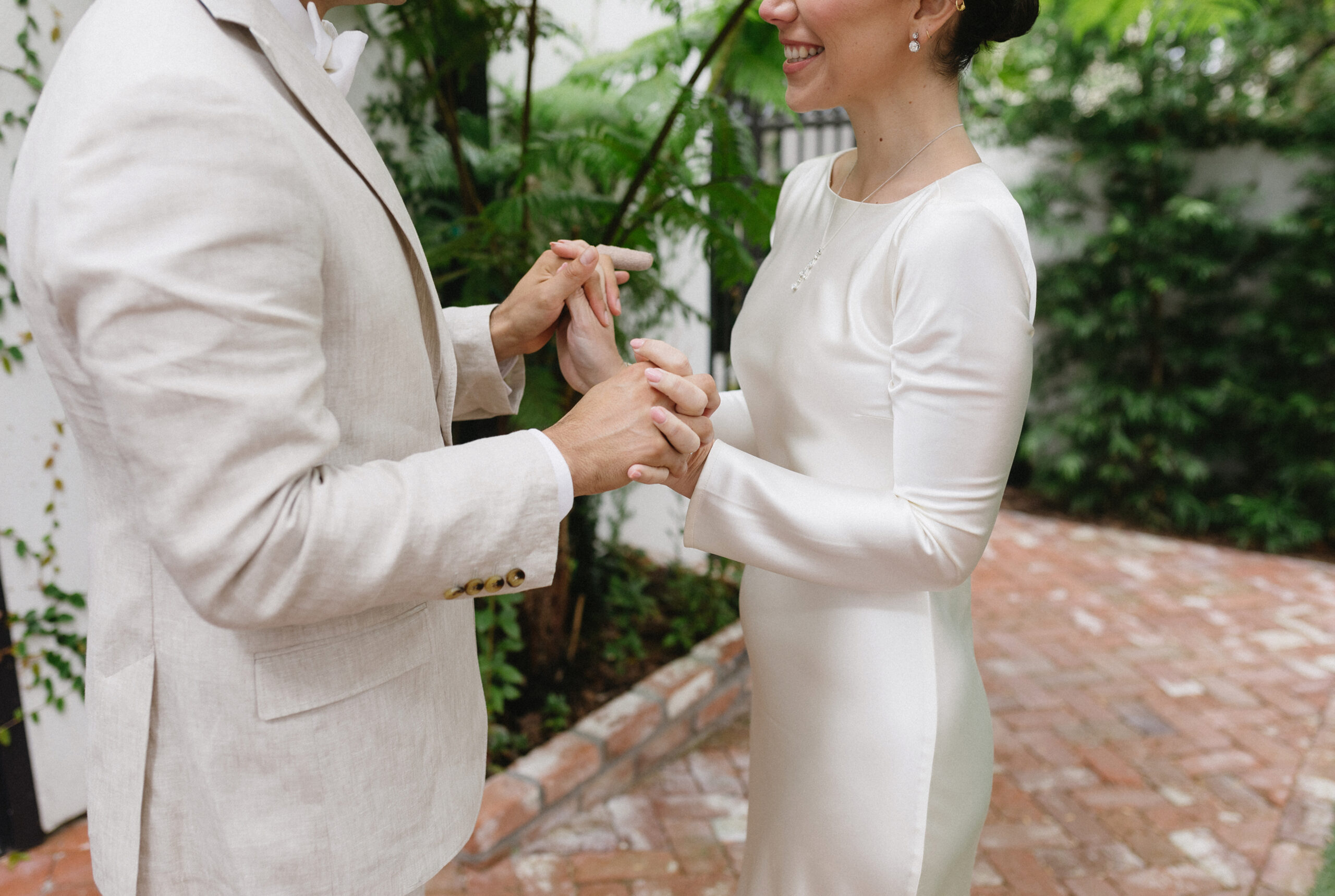Bride and groom holding hands and smiling at each other during an intimate ceremony in a garden courtyard.