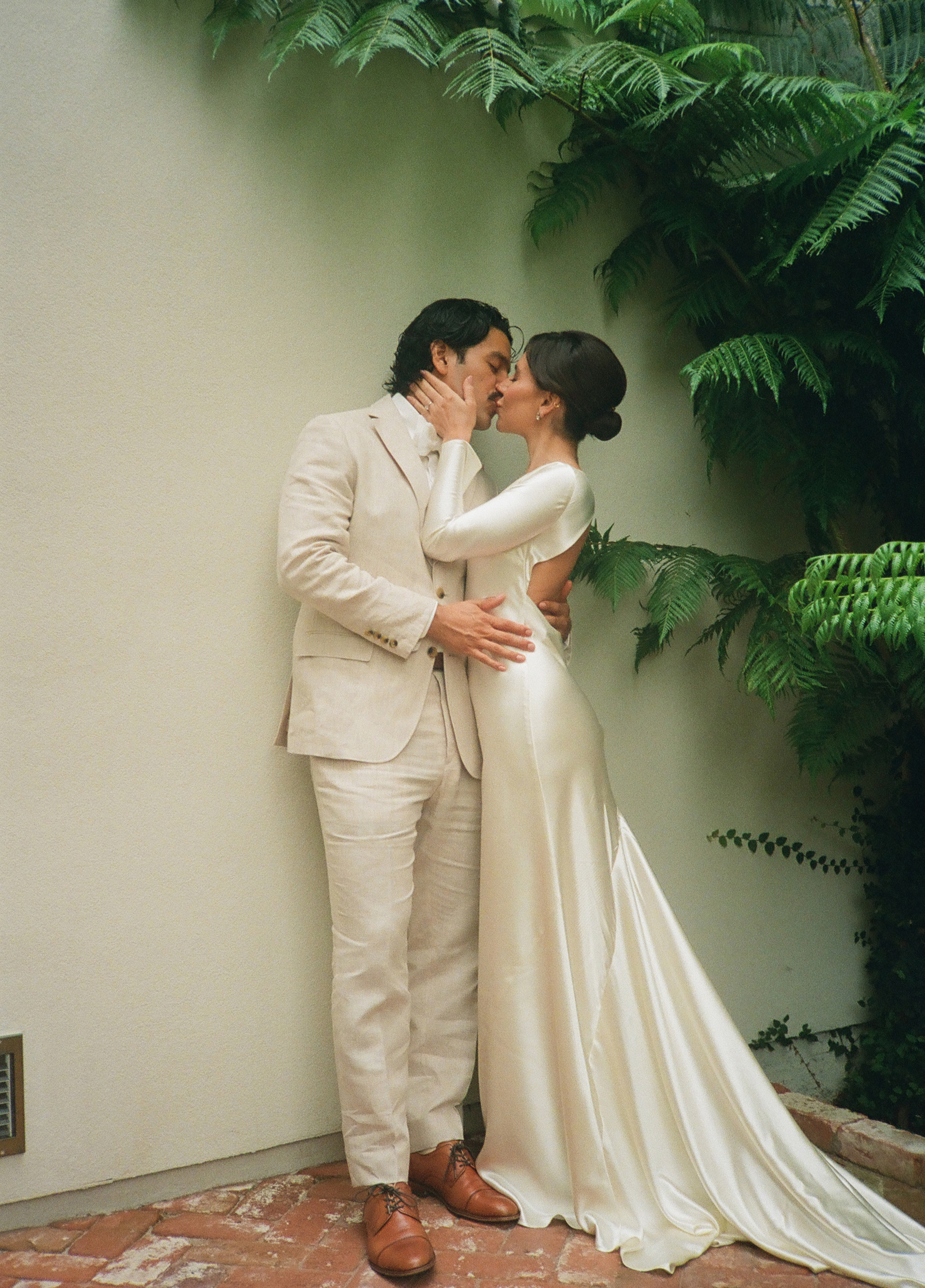 Bride and groom sharing a kiss in a garden courtyard, dressed in light-colored attire with lush greenery framing the scene.