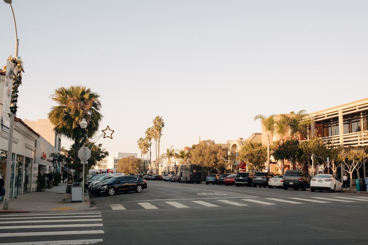HelloHorizon ORLI.Photos 34 Palm tree–lined street in a coastal town at golden hour with parked cars, crosswalks, and low-rise shops and buildings along both sides of the road.
