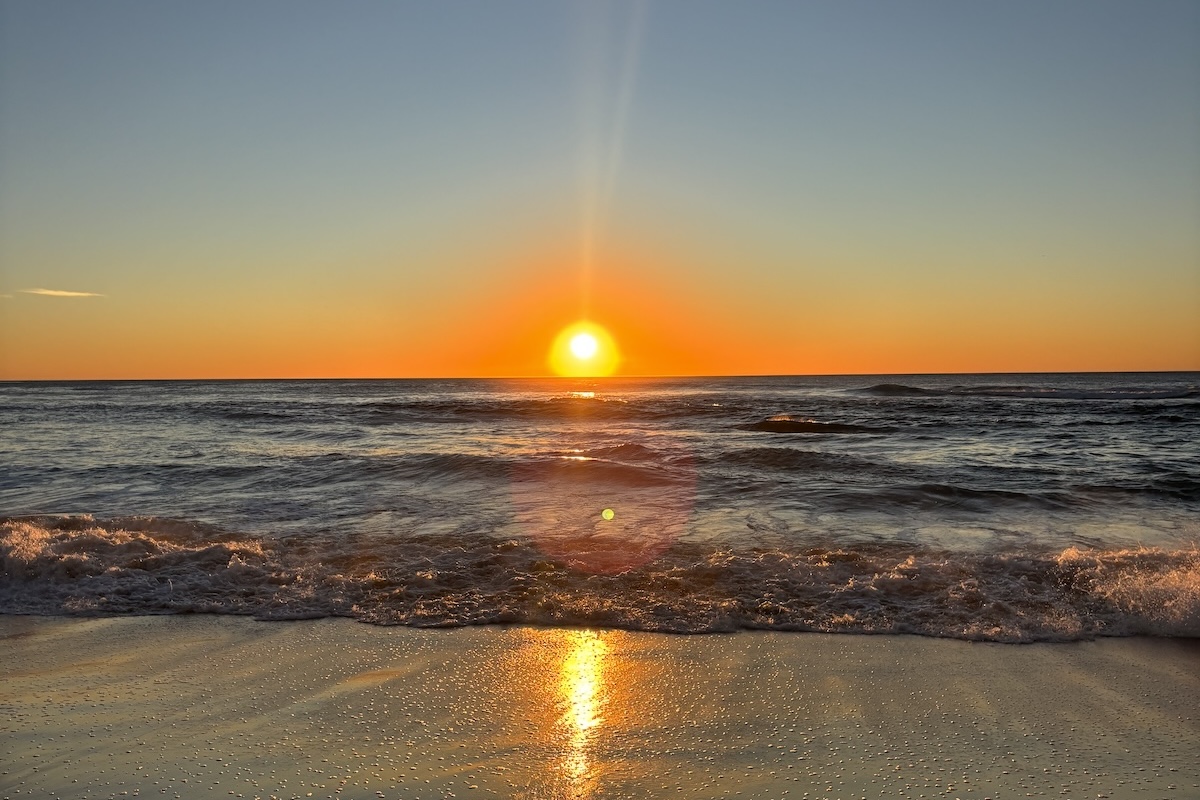 Bright orange sunset over the ocean with waves rolling onto the sandy shore and sunlight reflecting across the water and wet sand.