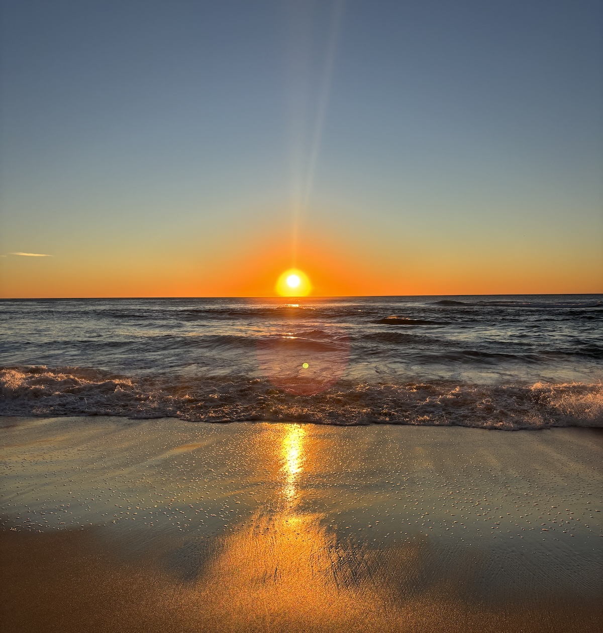 Bright orange sunset over the ocean with waves rolling onto the sandy shore and sunlight reflecting across the water and wet sand.