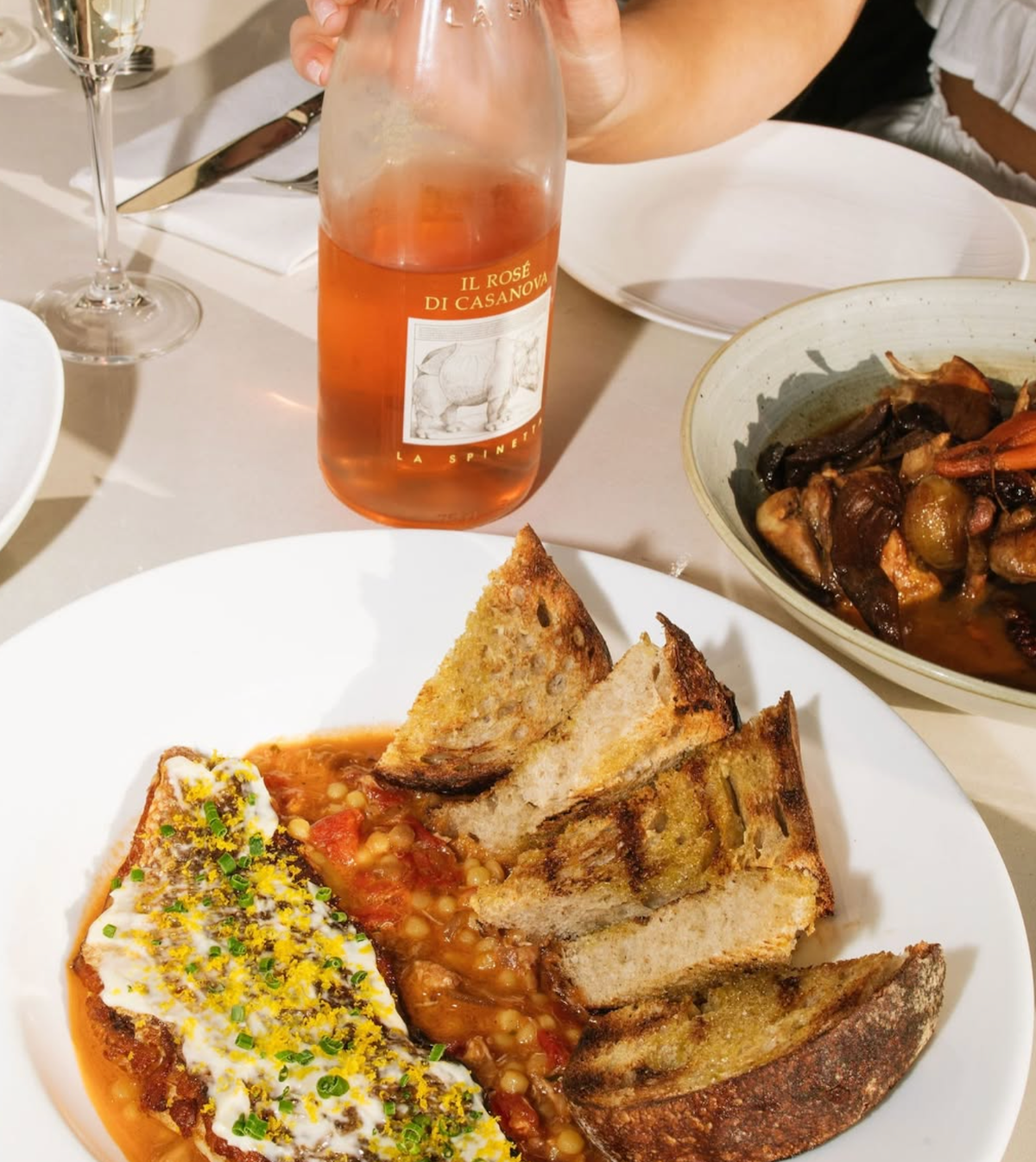 A restaurant table with a plate of toasted bread and a chickpea-based dish topped with yogurt and herbs, alongside a bottle of rosé wine and other dishes.