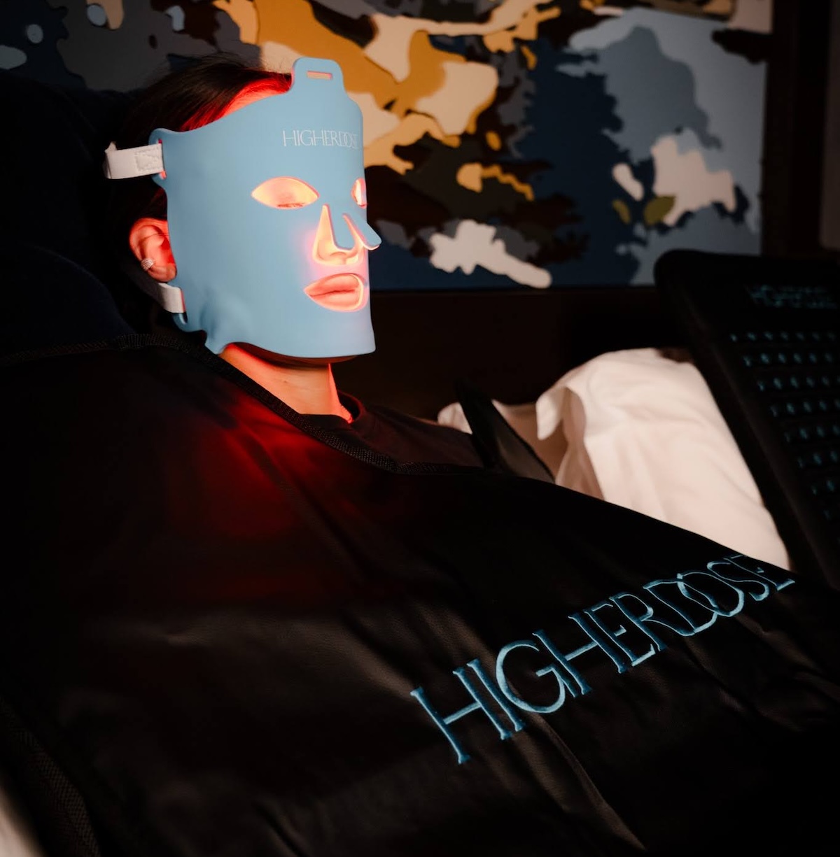 Person reclining while wearing a glowing LED facial mask, wrapped in a blanket in a dimly lit room.