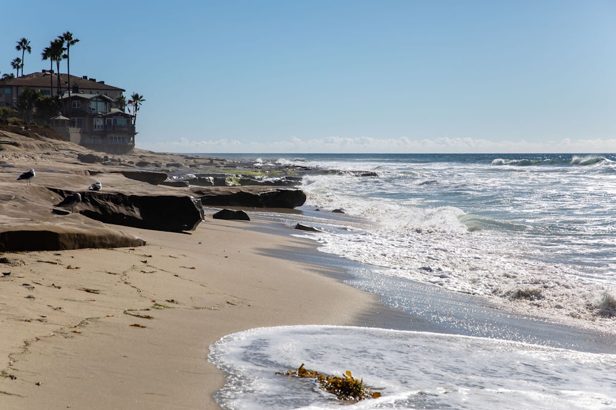 Sandy beach with gentle waves washing ashore beside rocky coastal formations, with oceanfront homes and palm trees in the background under a clear blue sky.