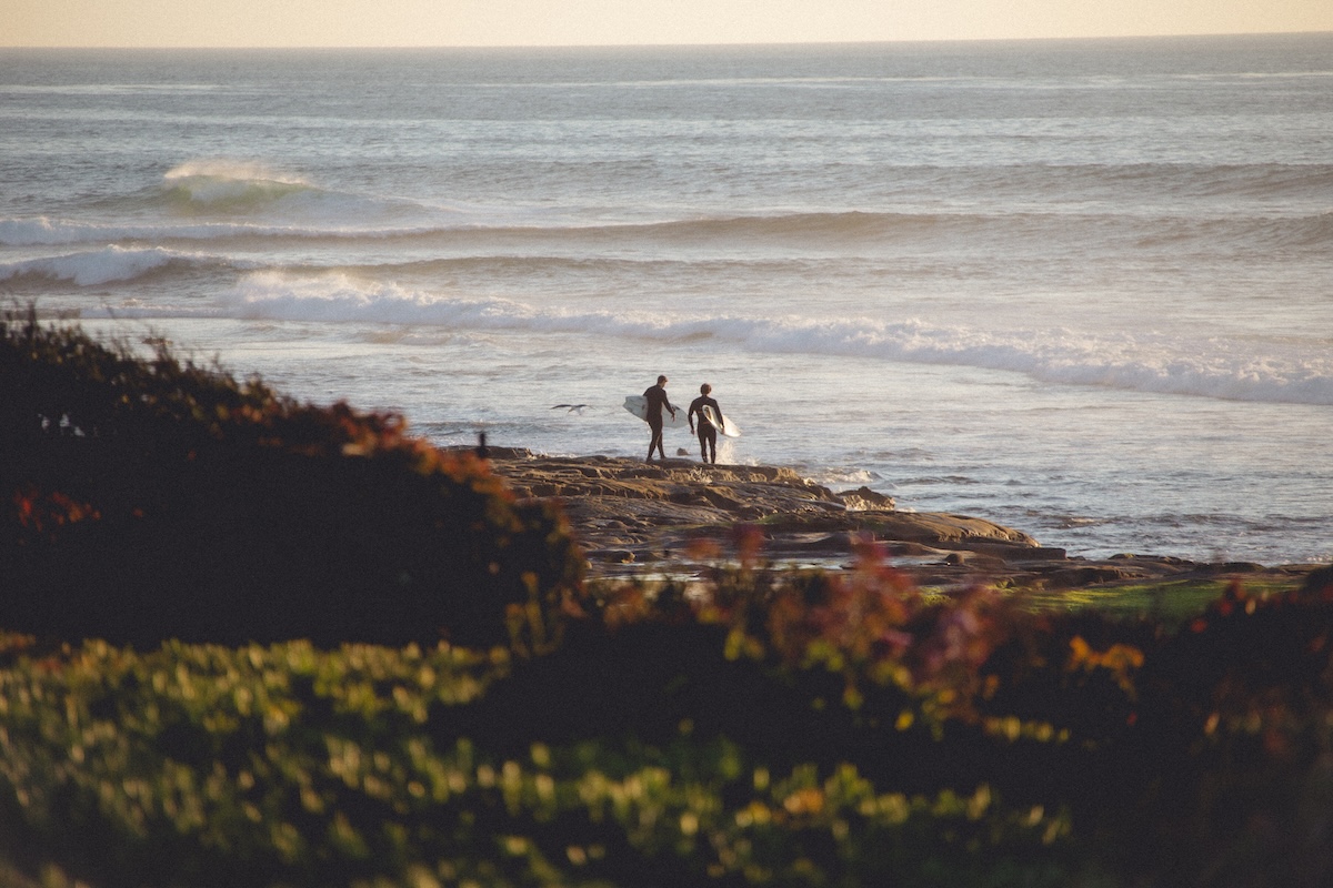 Two surfers carrying boards walk across a rocky shoreline beside rolling ocean waves in warm evening light.