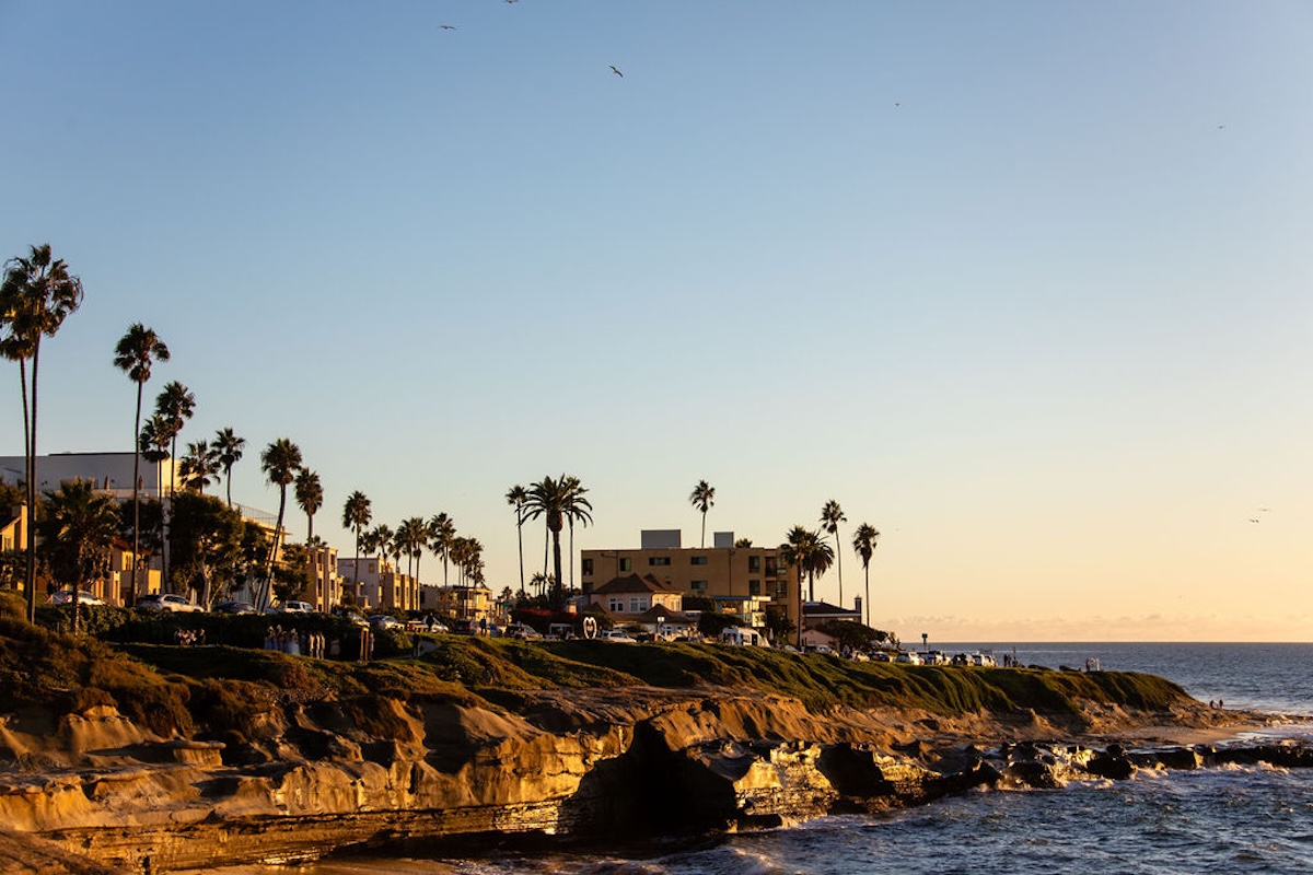 Coastal cliffs and ocean shoreline at sunset, with palm trees, buildings, and a clear sky overlooking the water.