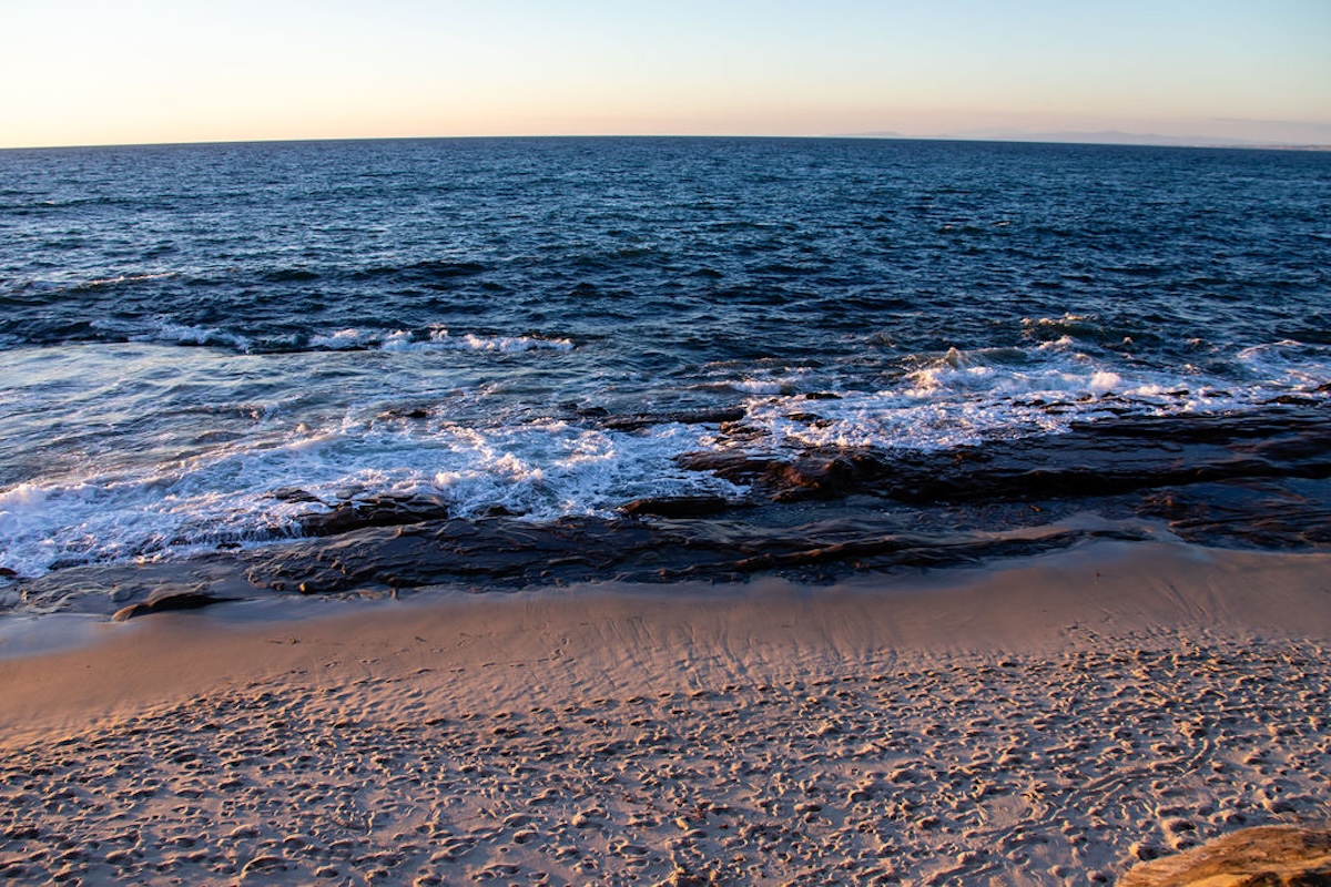 Ocean waves breaking over rocky shoreline beside a sandy beach, with open water stretching to the horizon under a clear sky.