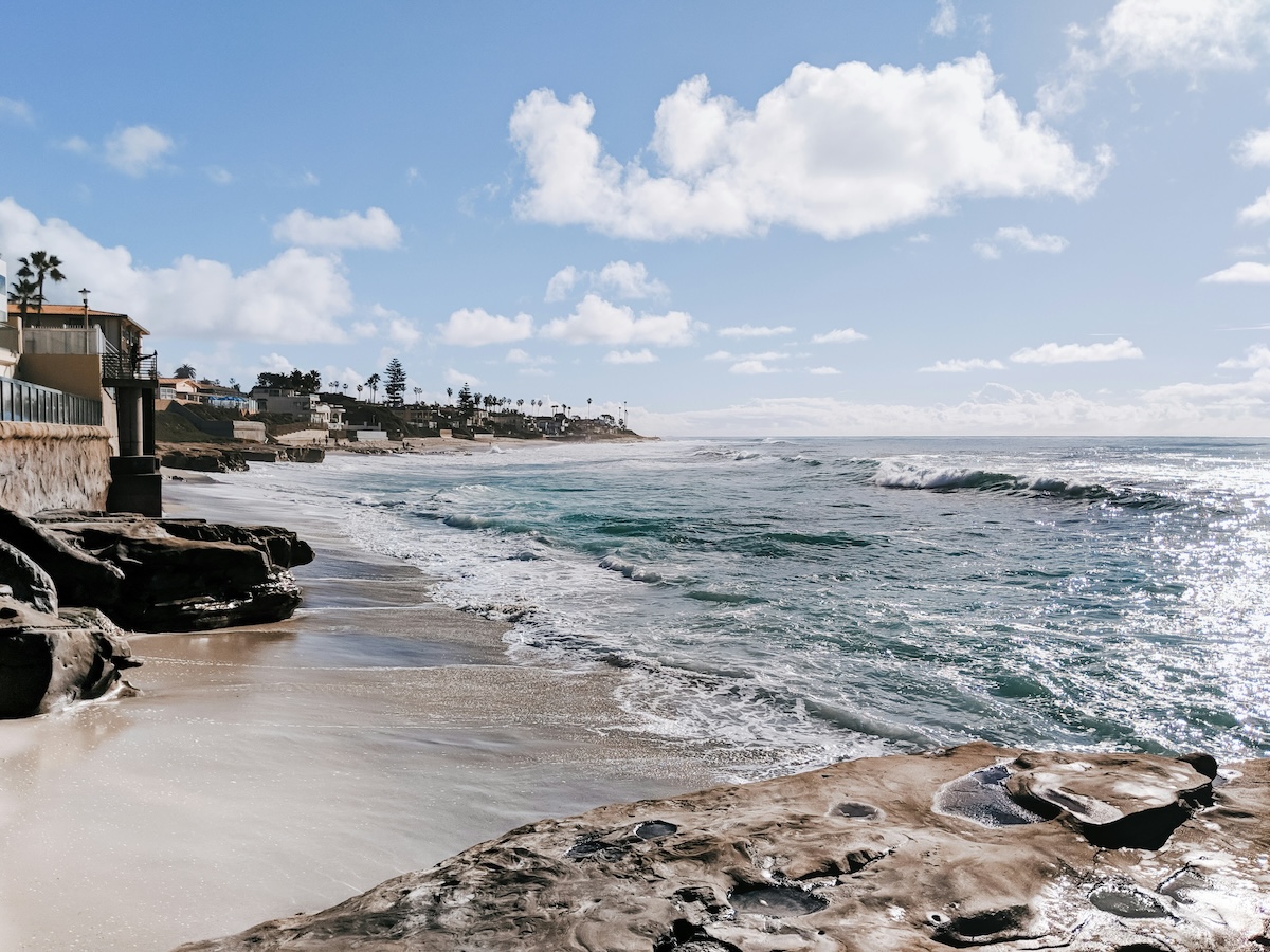 Waves wash onto a sandy beach bordered by smooth rock formations beneath a bright, partly cloudy sky.