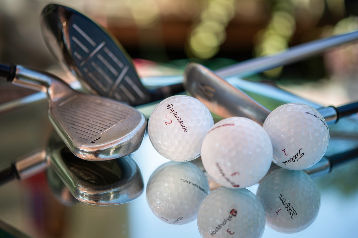 Several golf balls and irons rest on a reflective glass surface with a softly blurred background.