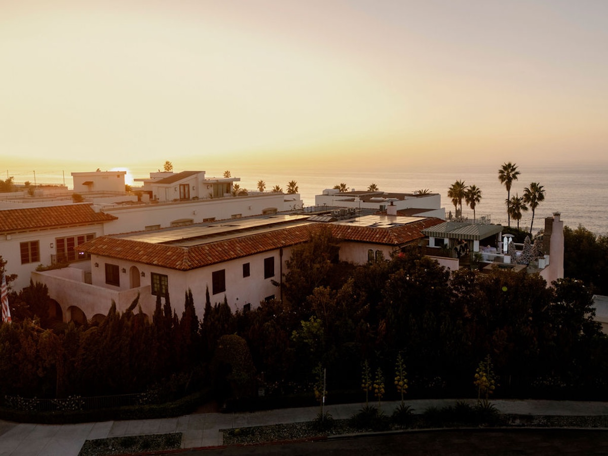 Golden sunset light illuminates Mediterranean-style homes and palm trees overlooking the ocean.