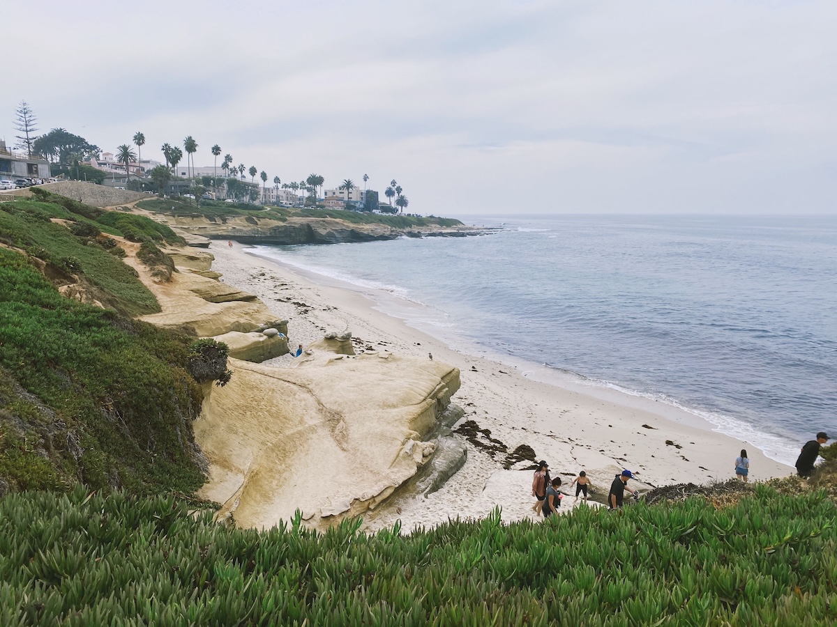 Elevated view of a sandy beach bordered by rocky coastal bluffs and shoreline vegetation, with calm ocean water, scattered beachgoers, and palm trees in the distance under a cloudy sky.